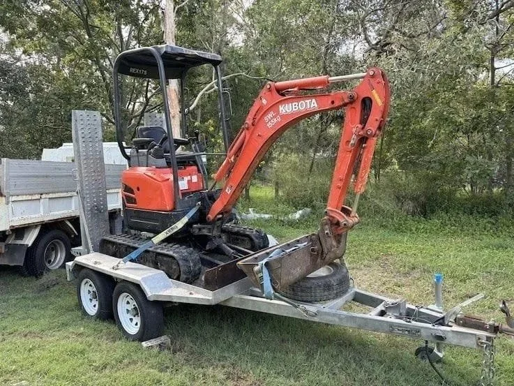A mini excavator on a trailer hitched to a pickup truck in an outdoor area with trees and grass.