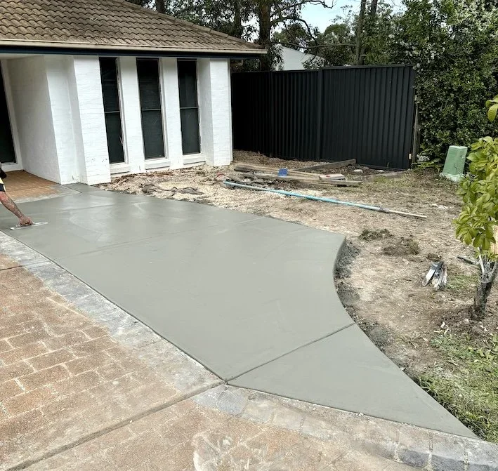 Construction site with a small excavator working near a large pile of dirt and a moving truck, with trees and a building in the background.