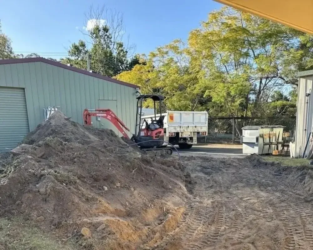 Construction site with a small excavator working near a large pile of dirt and a moving truck, with trees and a building in the background.