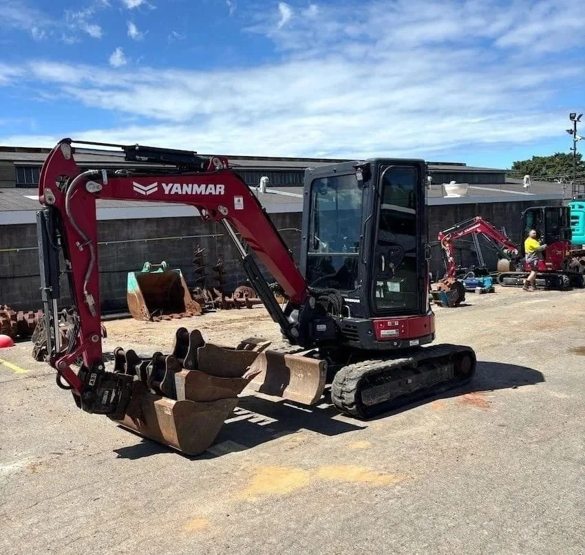 A small Yanmar excavator with a red arm and black body, sitting on a gravel lot, with other construction equipment and workers in the background.