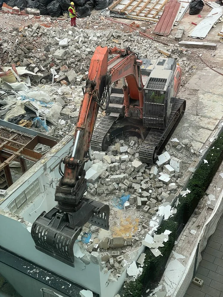 A construction site with a large excavator demolishing a building, rubble and debris scattered around, two construction workers in safety vests and helmets monitoring the work.