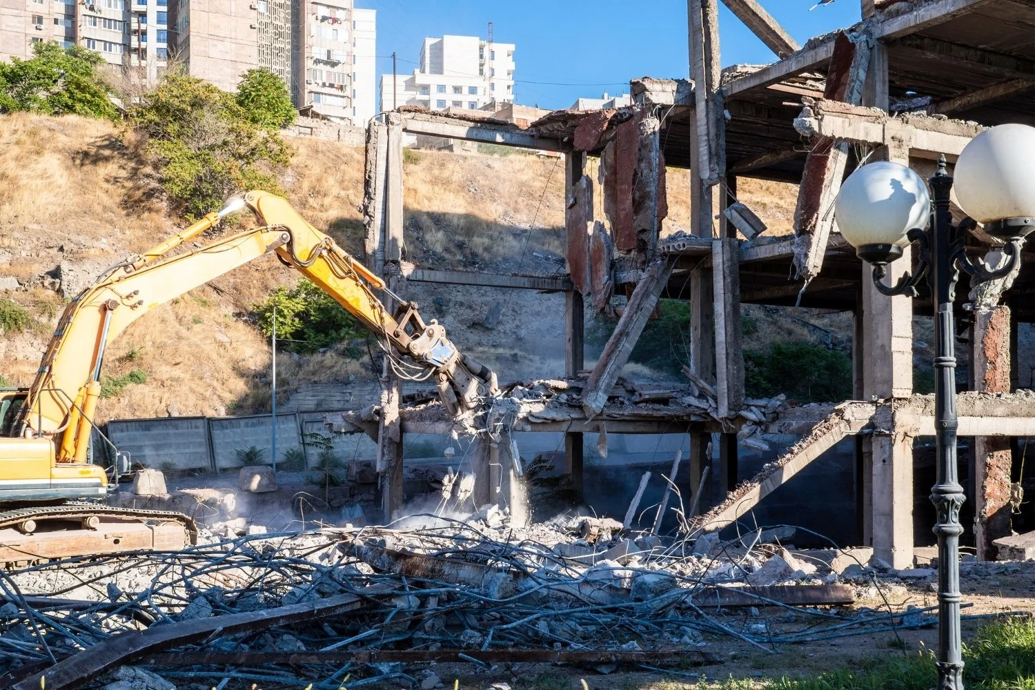 Demolition of a two-story building with exposed concrete and steel beams, rubble and debris on the ground, an excavator, and a lamppost in the foreground; in the background, tall apartment buildings on a hillside and a clear blue sky.