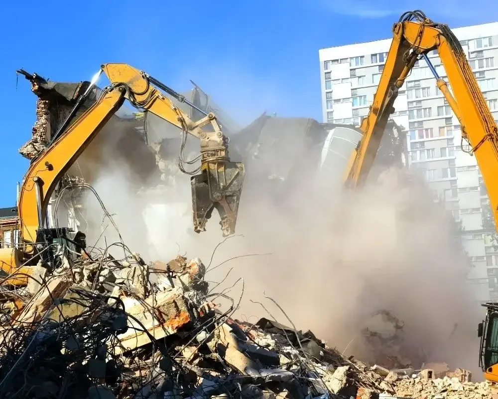 Two excavators demolishing a building, with dust and debris in the air and rubble on the ground.