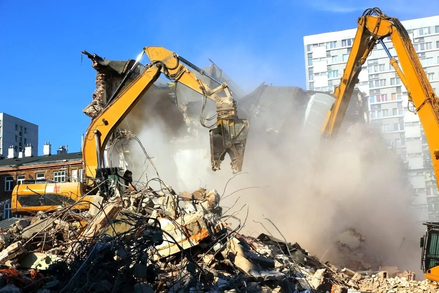 Two yellow excavators demolishing a building, with debris and dust in the air, and urban buildings in the background.
