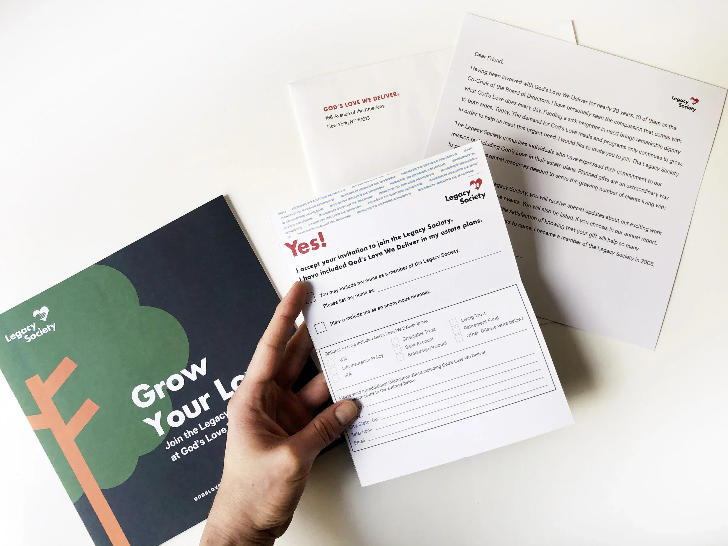 Hand holding a paper with a charity donation form from Legacy Society, on a white desk with other informational papers and a brochure for the organization.
