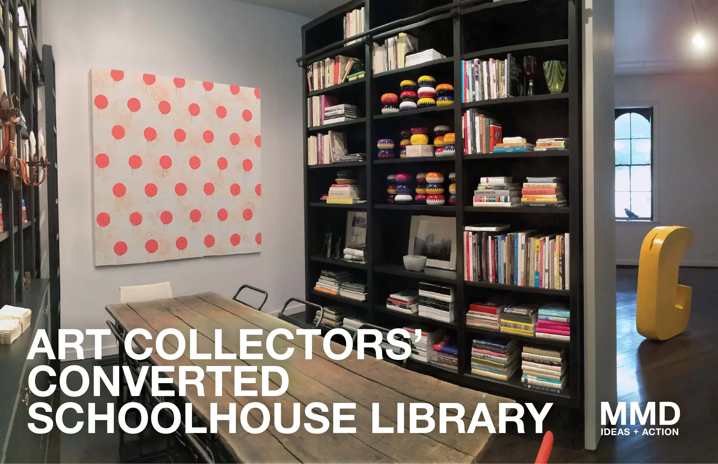 Interior of a converted schoolhouse library with a large black bookshelf filled with books, art pieces, and decorative items, including colorful bowls. There is a wooden table with chairs and a prominent pink polka dot artwork on the wall. The image 