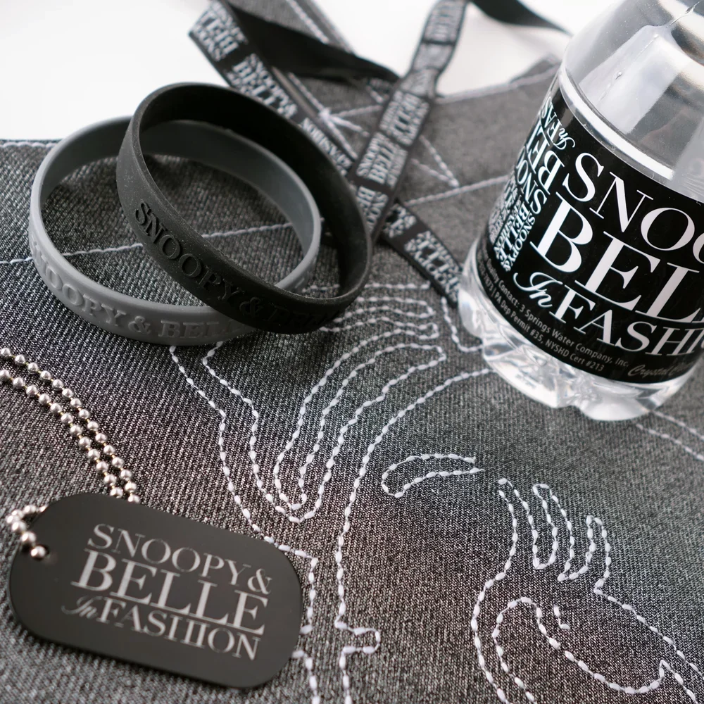Black and gray silicone wristbands, a plastic water bottle from Snoopy & Belle's Fantasy, and a black dog tag necklace from Snoopy & Belle's Fantasy on a denim fabric with white stitched embroidery.