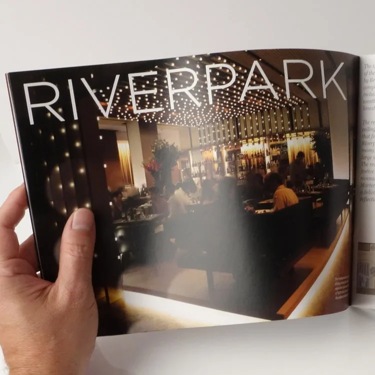 Interior of a restaurant or lounge named Riverpark, with dining tables, people sitting, warm lighting, and decorative elements.
