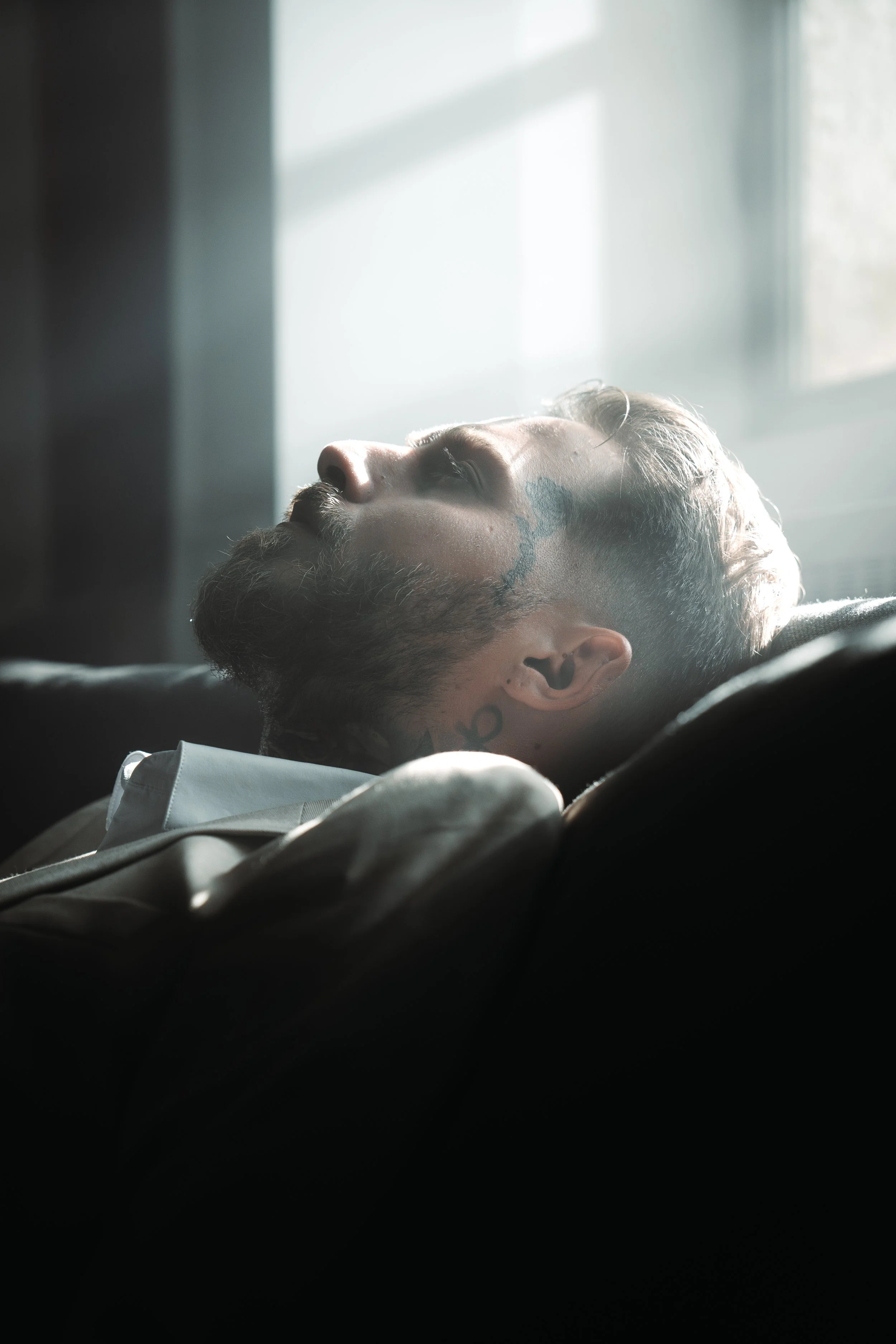 Un homme avec une barbe et des tatouages, dormant ou se reposant, sous un éclairage naturel provenant d'une fenêtre.