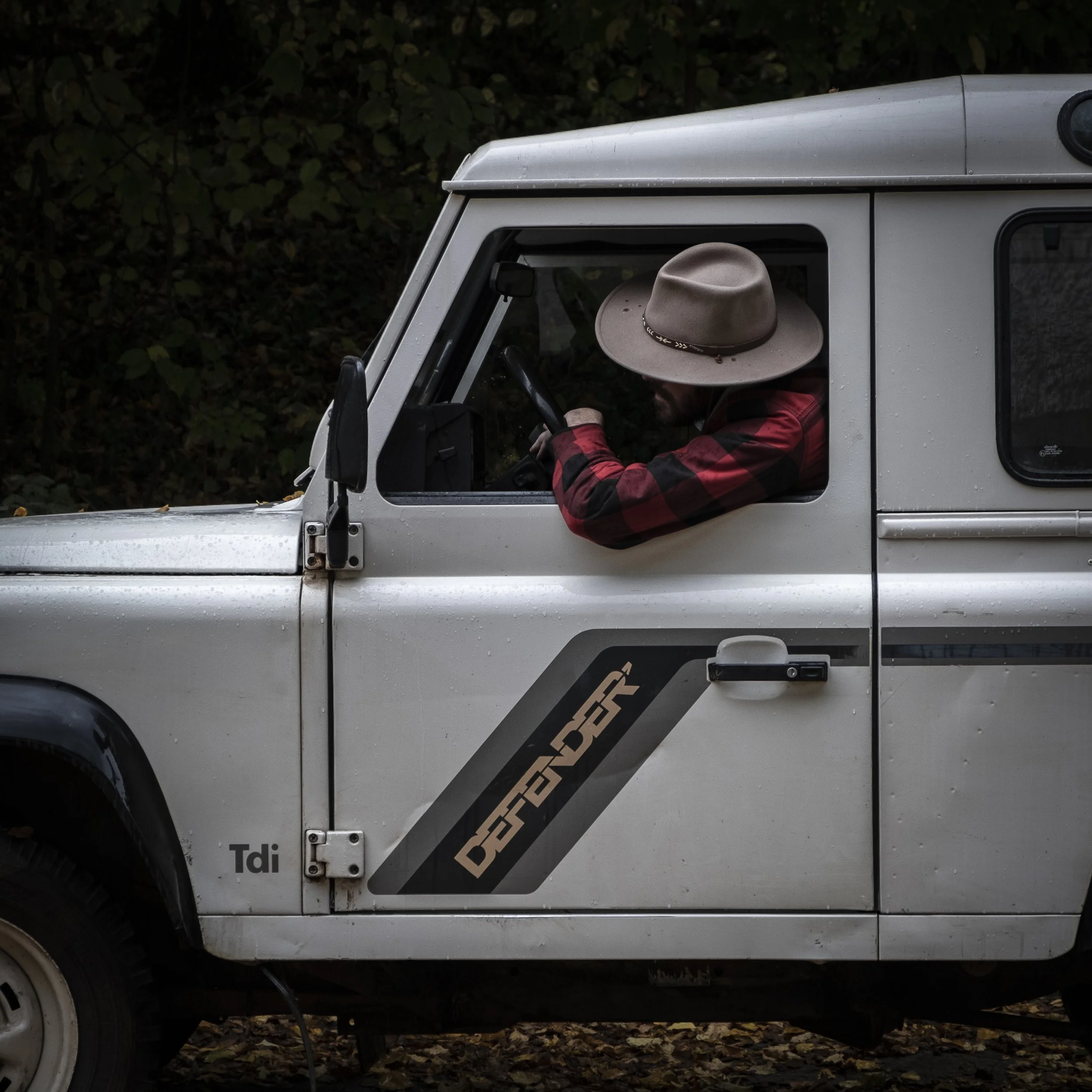 Un homme portant un chapeau beige et un manteau à carreaux rouges et noirs, assis dans le véhicule Land Rover, en train de regarder le volant.