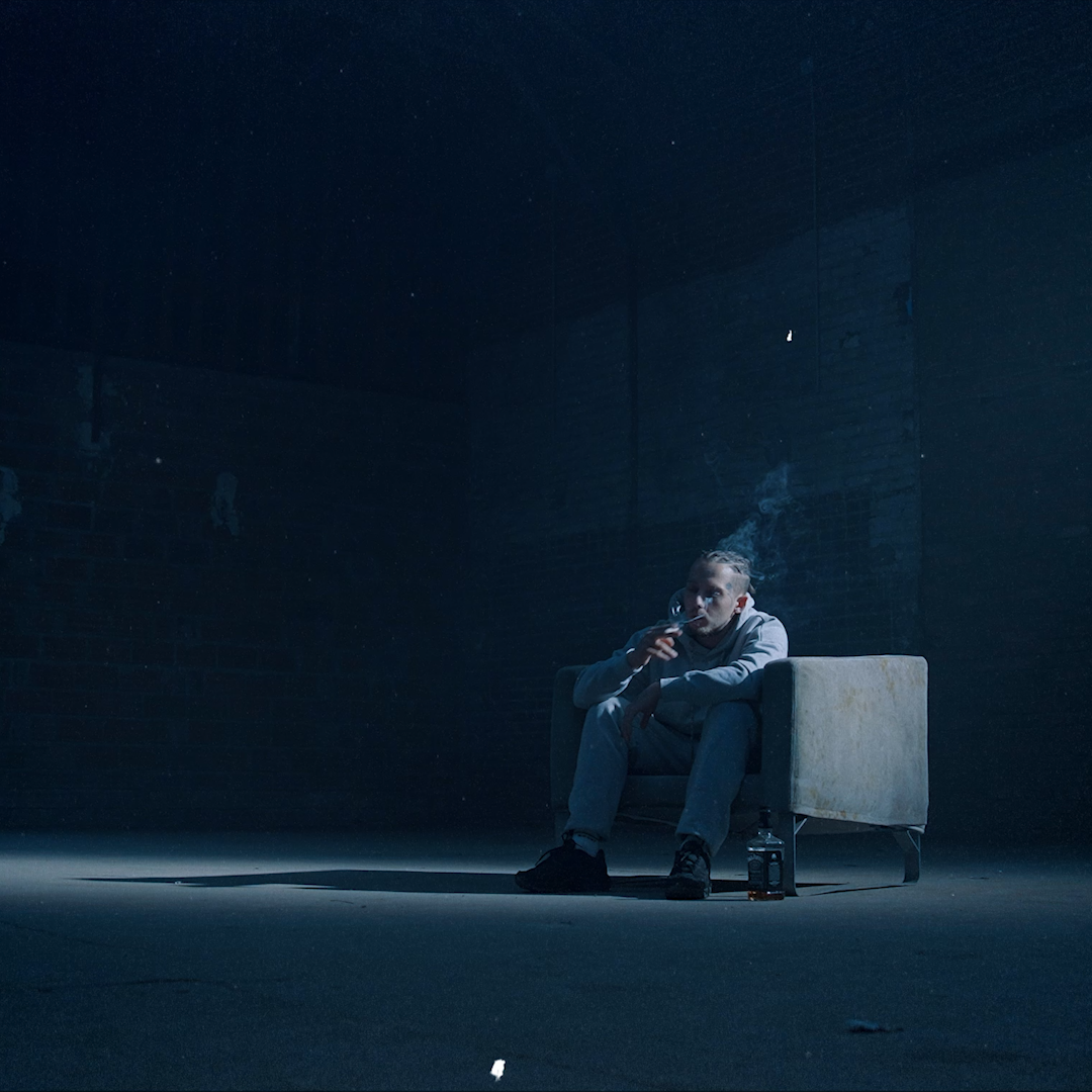 Homme assis sur un canapé en train de fumer dans une pièce sombre, avec une bouteille d'alcool posée au sol à côté.