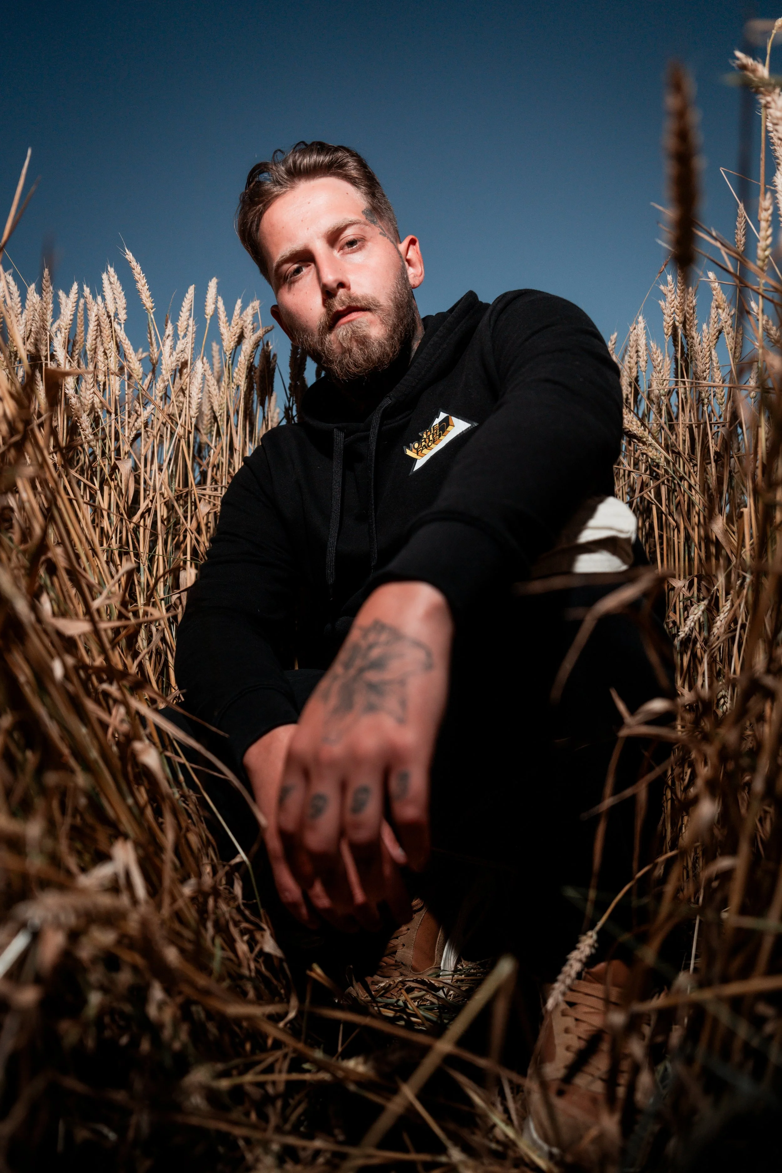 Un homme assis dans un champ de blé, portant un sweat noir, avec un tatouage sur la main, sous un ciel bleu clair.
