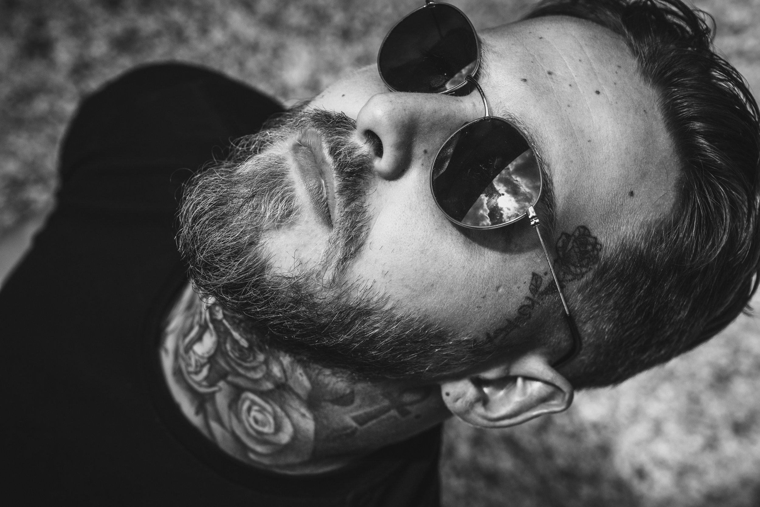 Un homme avec des tatouages, des lunettes de soleil et une barbe, vue de haut, en noir et blanc.