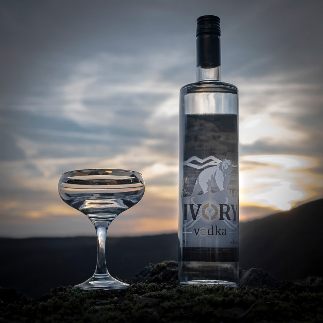Une bouteille de vodka Ivory et un verre à cocktail, tous deux placés sur une surface naturelle, avec un coucher de soleil en arrière-plan dans un paysage montagnard.