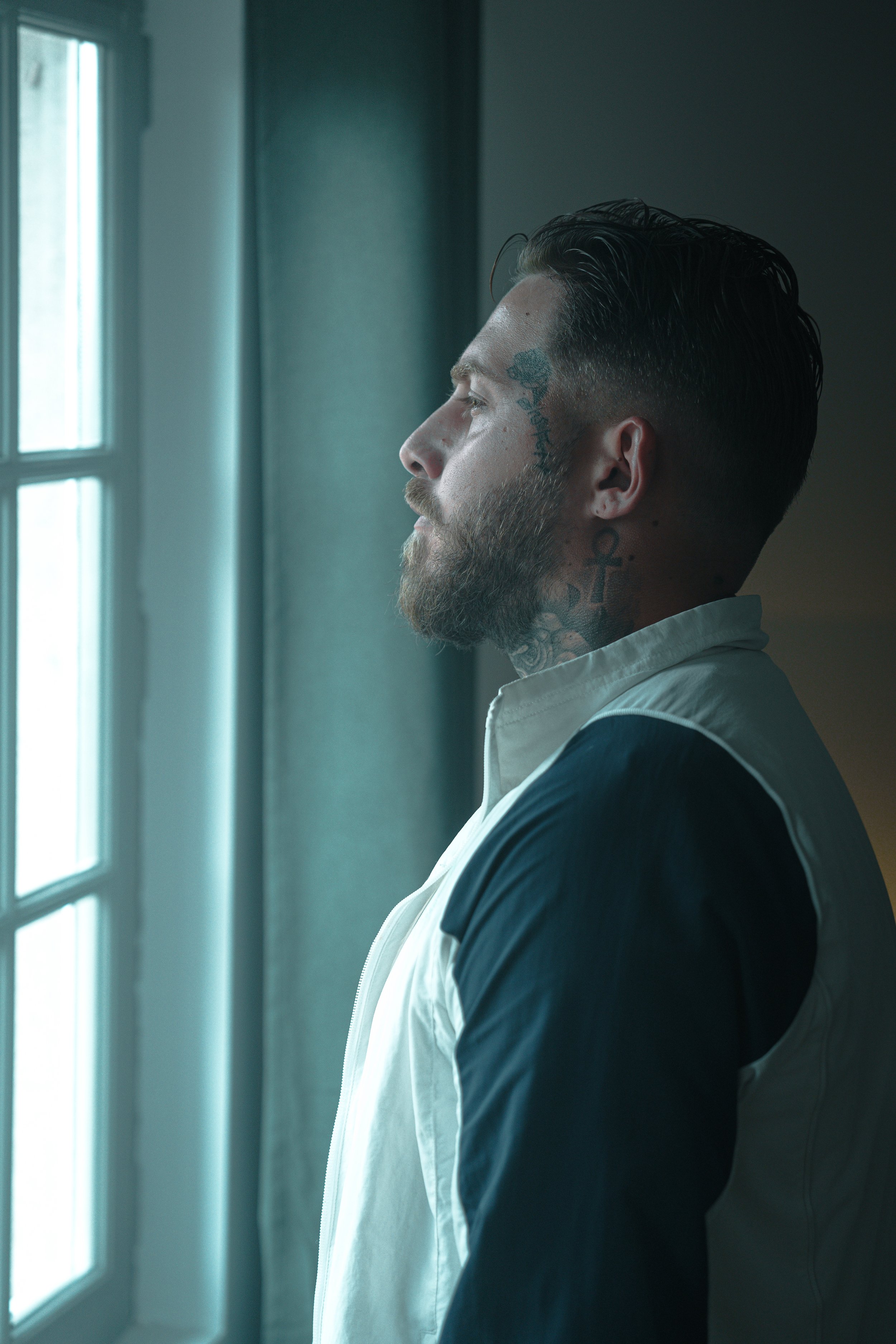Un homme avec une barbe et des tatouages, regardant par une fenêtre, dans une pièce sombre