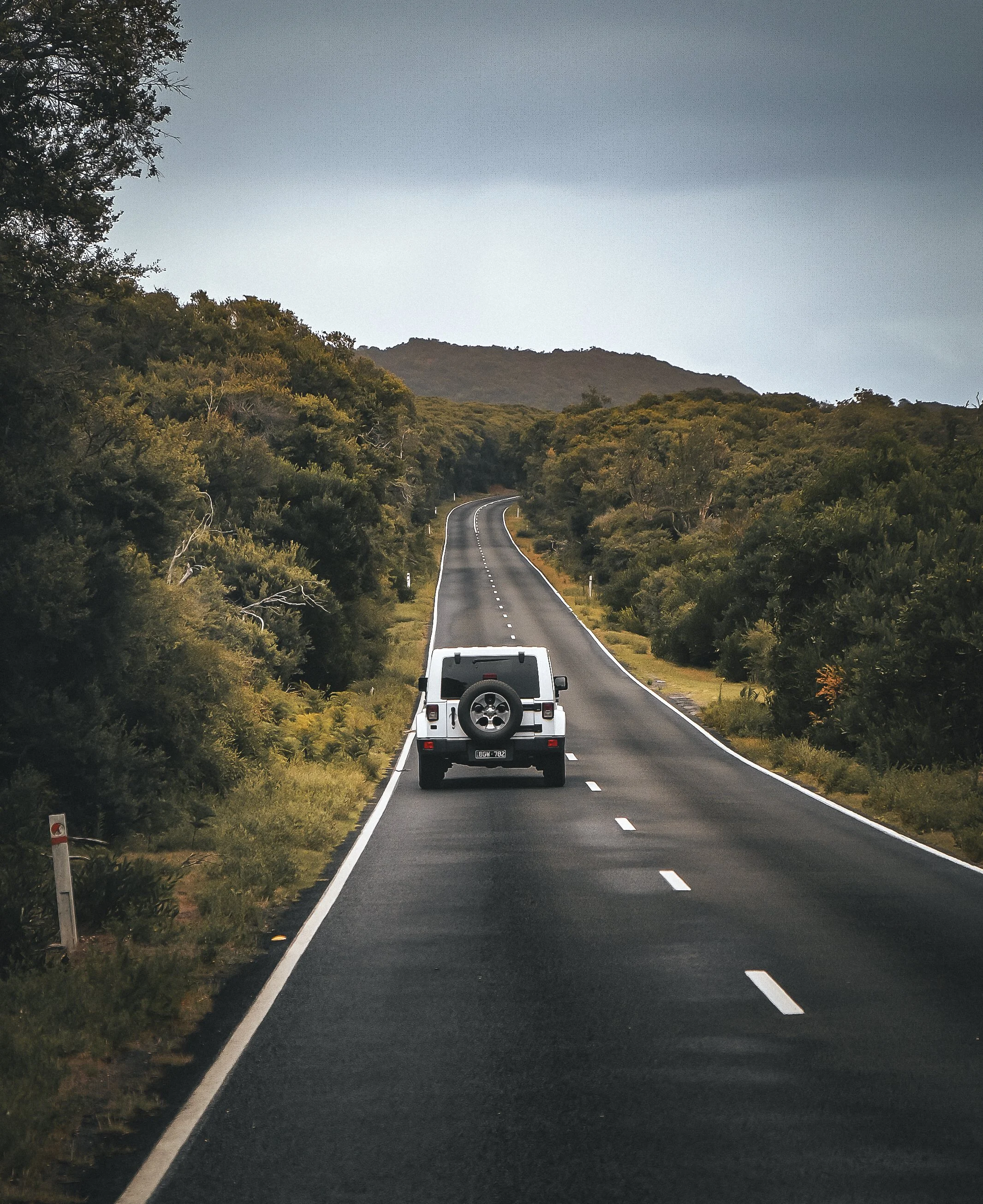 Voiture blanche sur une route de campagne sinueuse entourée de végétation dense et de collines sous un ciel nuageux.