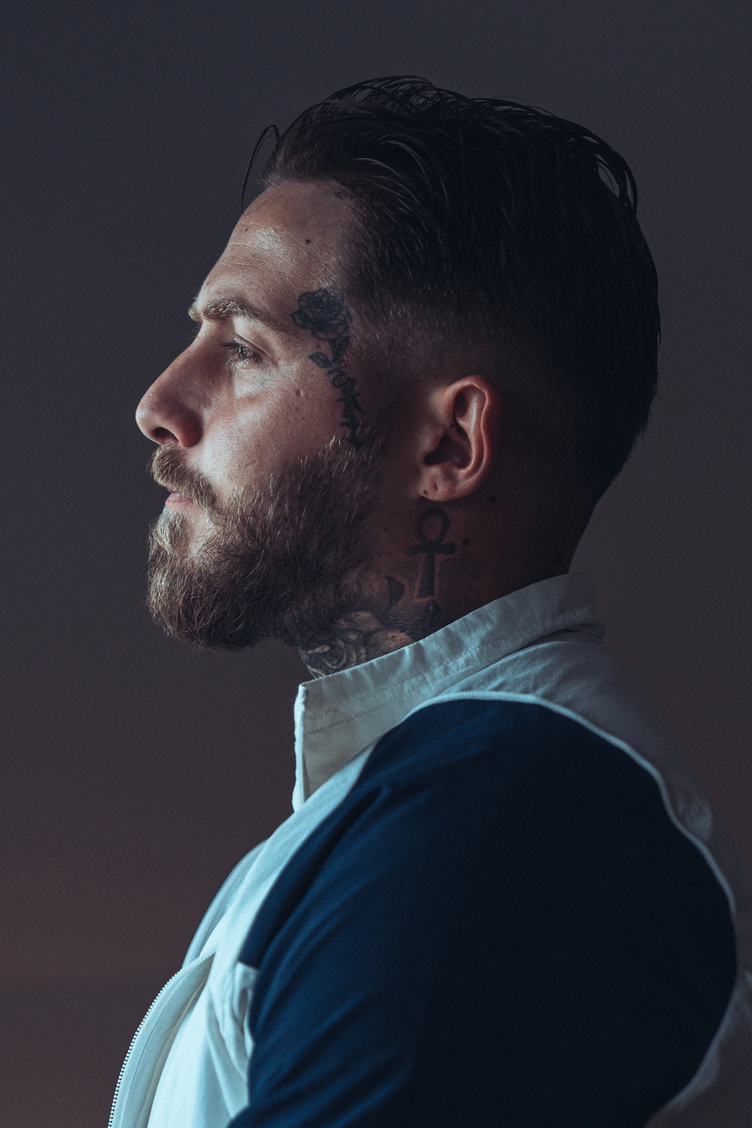 Profil d'un homme avec tatouages sur le visage et le cou, cheveux en arrière, portant une veste blanche et bluette, fond sombre.