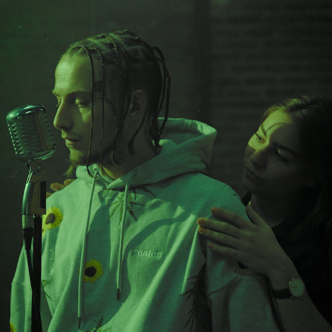 Deux personnes dans un studio d'enregistrement, une devant un microphone vintage, l'autre la touche à l'épaule, ambiance sombre avec lumière verte.