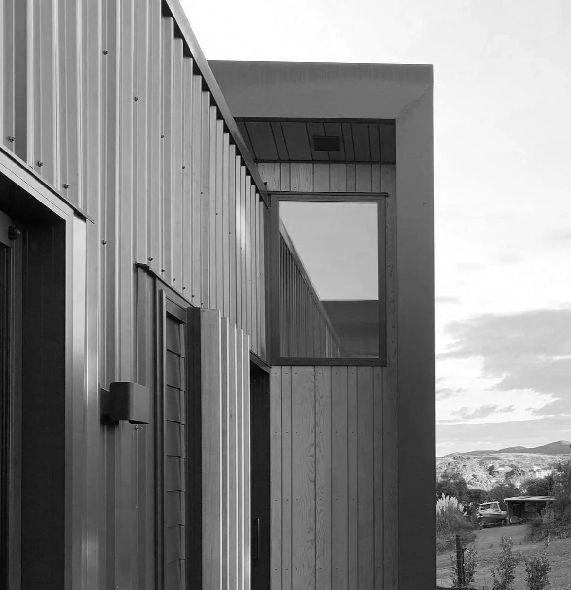 Black and white photo of a modern house with wooden exterior, large window, and a view of landscape with hills and sky in the background.
