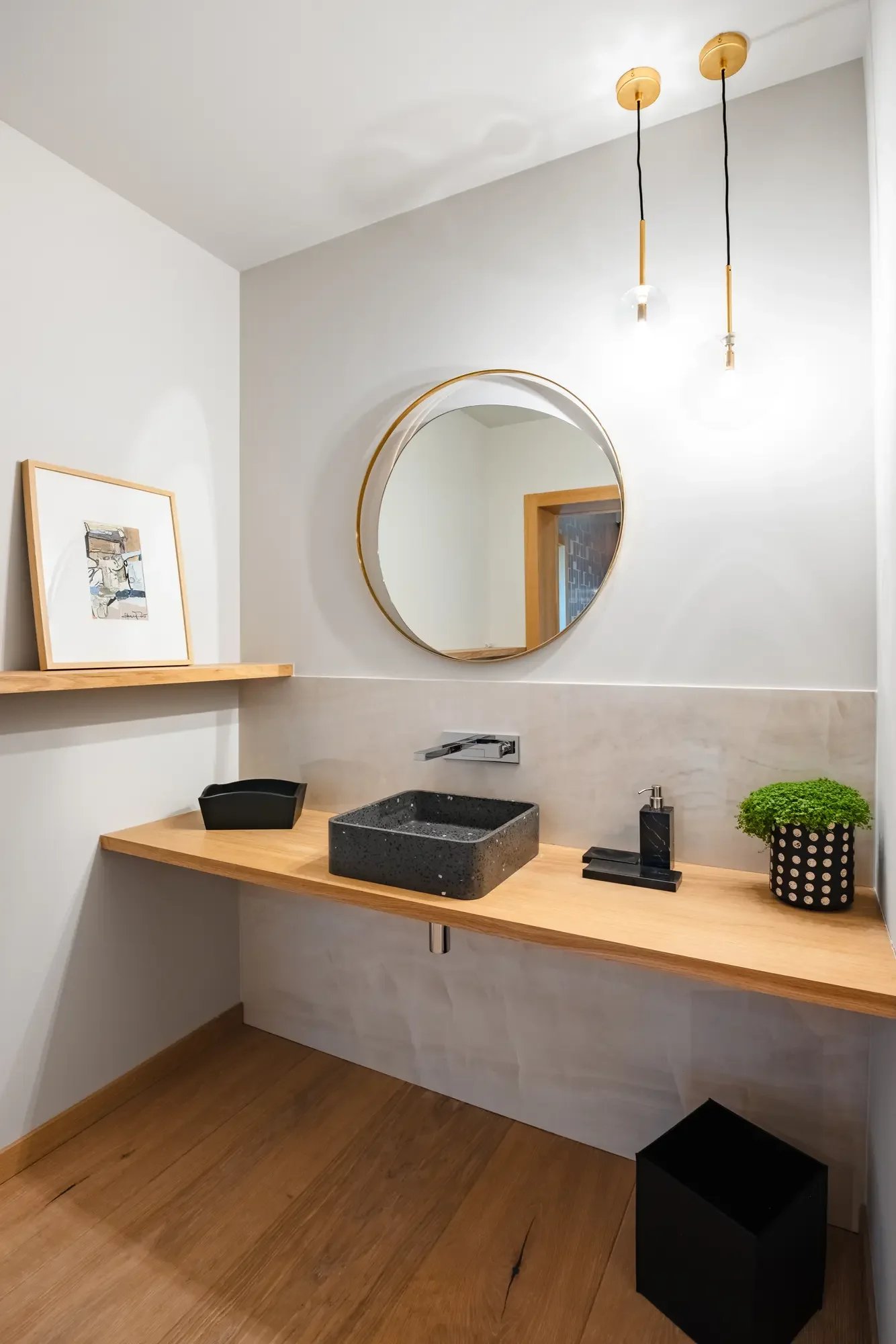 Salle de bain minimaliste avec un miroir rond, un lavabo carré en pierre noire sur un comptoir en bois, un évier mural, une plante verte, un porte savon noir, une étagère en bois, et deux suspensions au plafond.