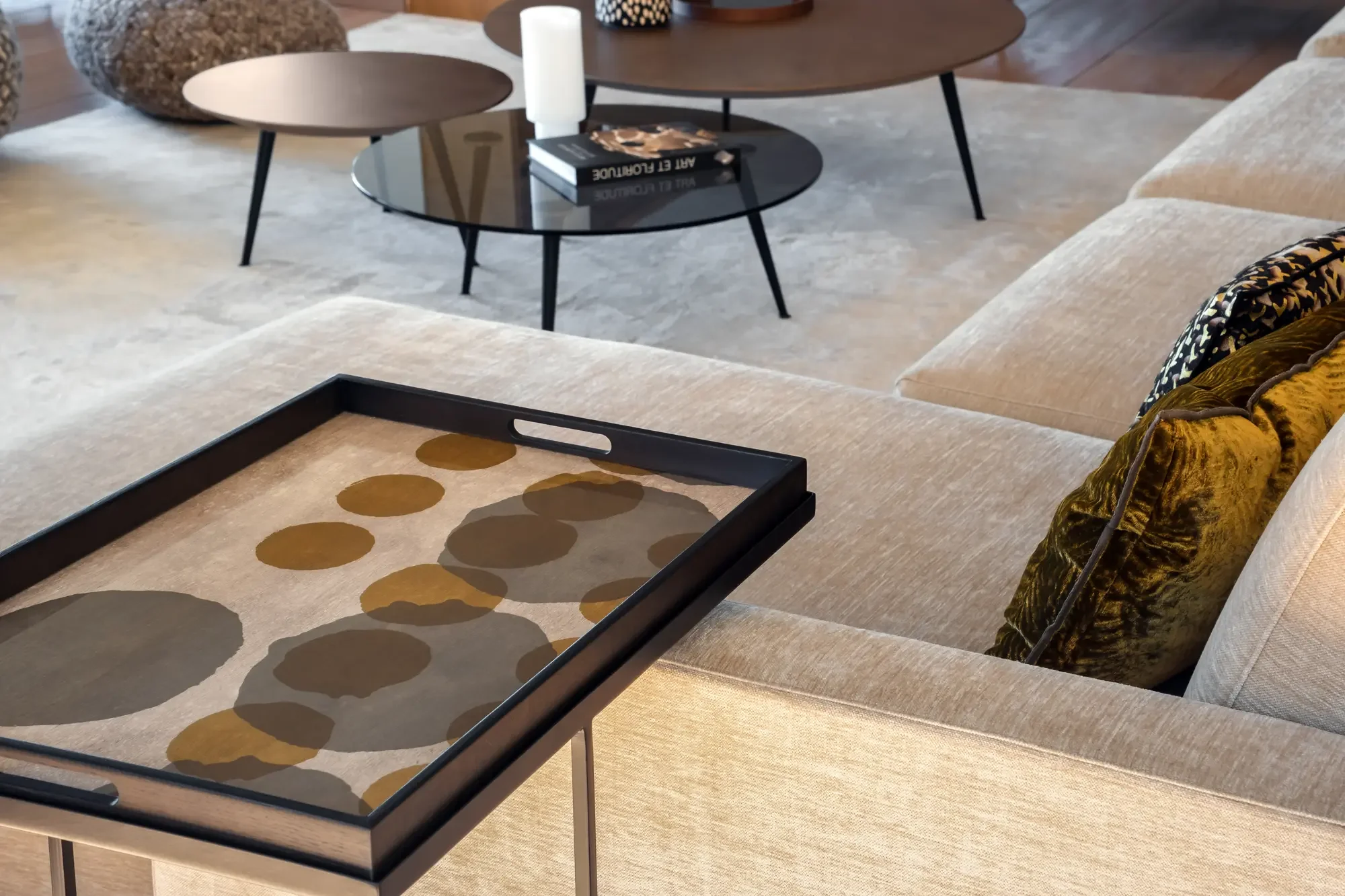 Un salon avec un canapé beige, des coussins en velours dorés et noirs, une table basse noire avec des livres, un plateau décoratif avec des cercles abstraits, et deux tables gigognes en bois foncé