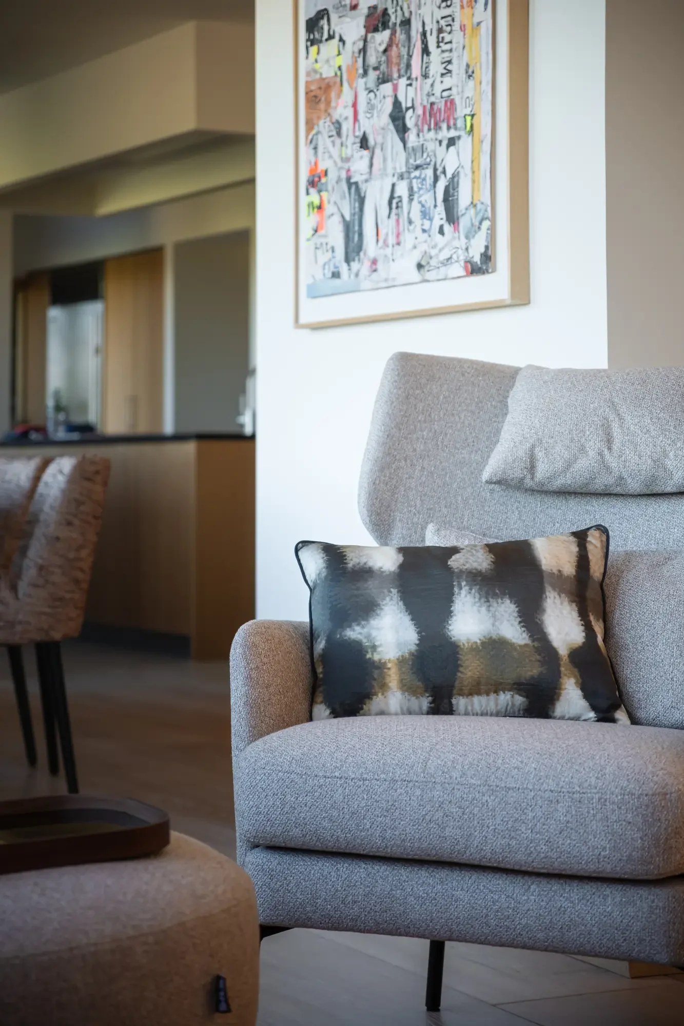 Un séjour intérieur avec un fauteuil gris, un coussin noir et blanc, une table basse en bois, une peinture moderne sur le mur et une cuisine en arrière-plan.