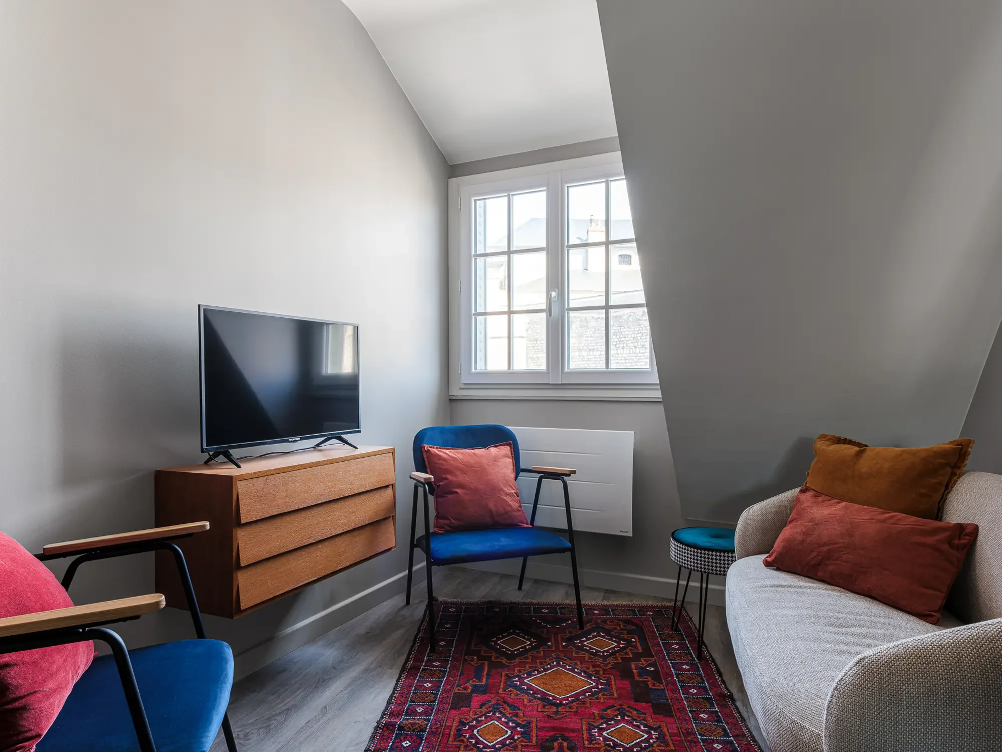 Salon avec fauteuils bleus et beige, canapé beige avec coussins marron et rouge, télévision sur un meuble en bois, tapis rouge avec motifs géométriques, fenêtre avec vue sur le toit, décoration minimaliste.