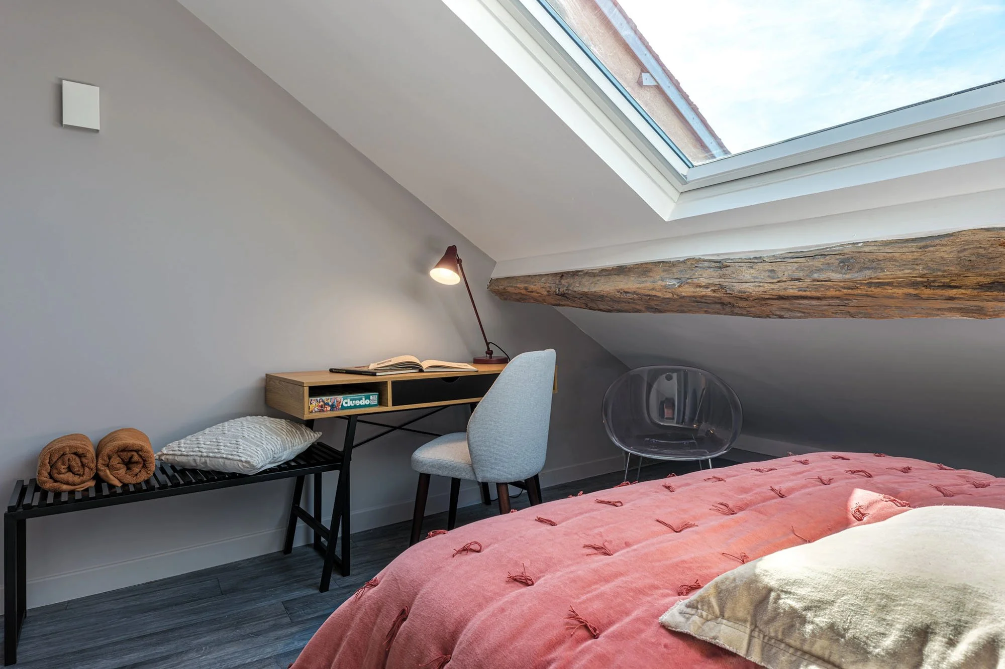 Une chambre dans un bureau mansardé avec un lit aux draps couchés, un bureau avec une lampe de lecture et un livre ouvert, un siège en plastique transparent, un fauteuil gris et une petite table en bois, un plafond avec une poutre en bois et un grand