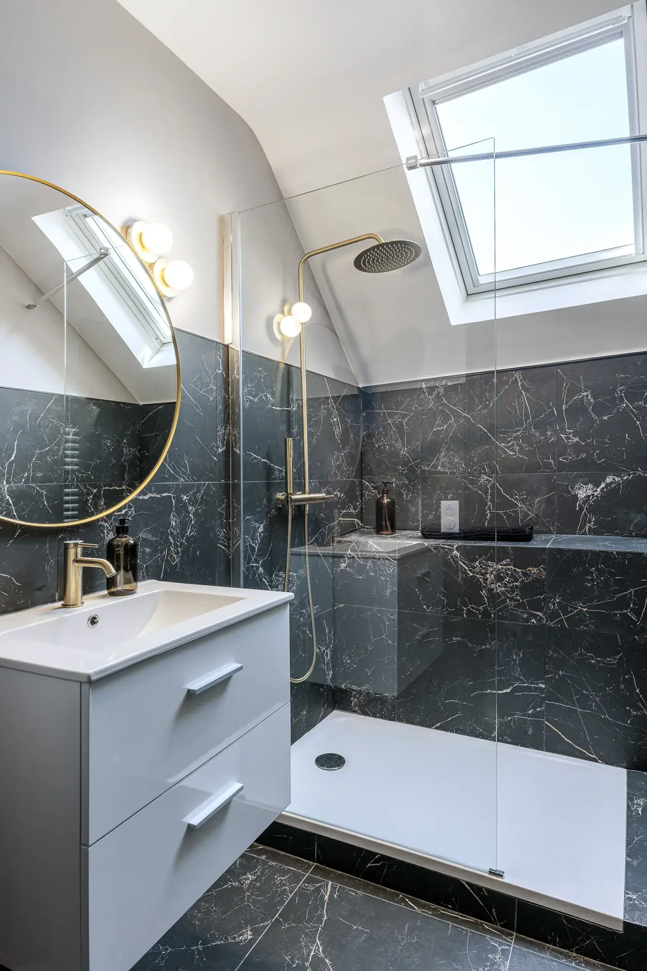 Salle de bain moderne avec murs en marbre noir, lavabo blanc avec meuble de rangement, miroir rond avec cadre doré, pommeau de douche noir, fenêtre de toit pour éclairage naturel, et sol en marbre noir