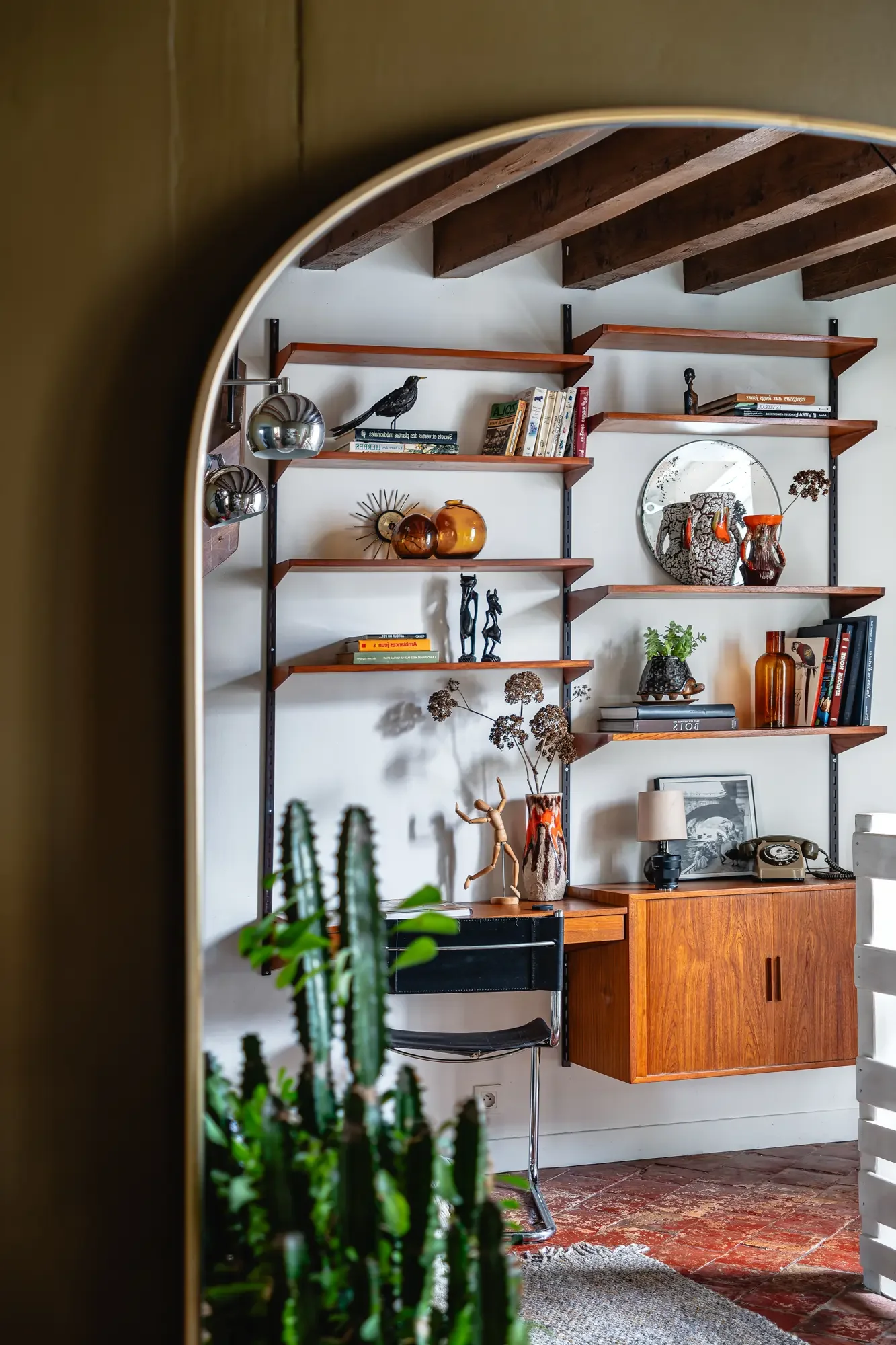 Intérieur d'une pièce avec une étagère en bois décorée de livres, vases et objets d'art, vue partielle à travers un miroir ou une ouverture arquée.