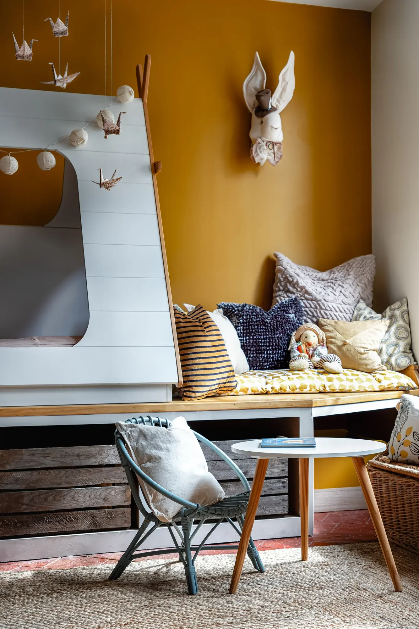 Coin salon avec espace de lecture, coussins, une figurine de poupée, et décorations murales telles qu'un lapin en tissu et des origamis d'oiseaux.