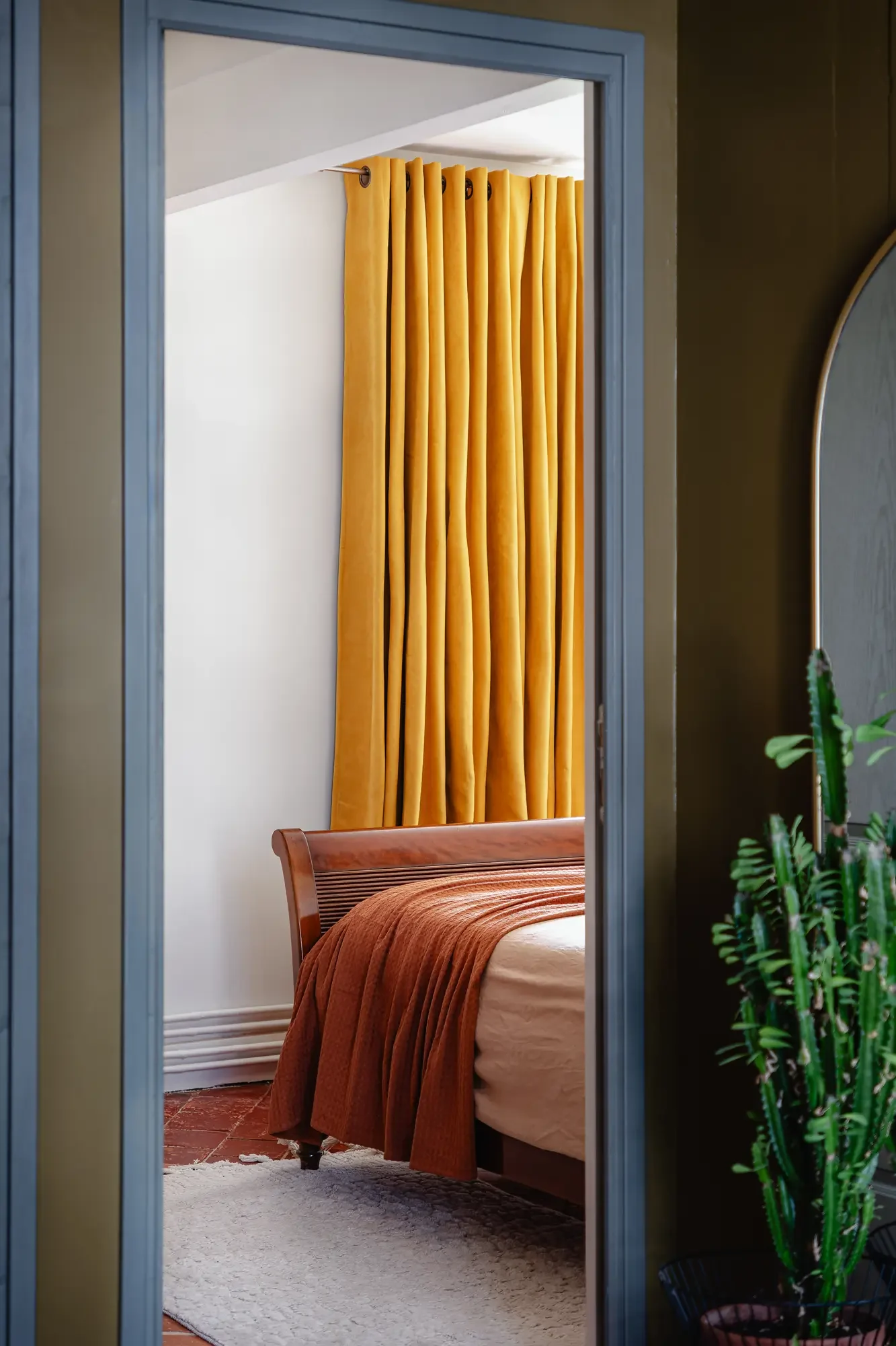 Une chambre vu à travers une porte, avec un lit en bois, des rideaux jaunes et un tapis blanc.