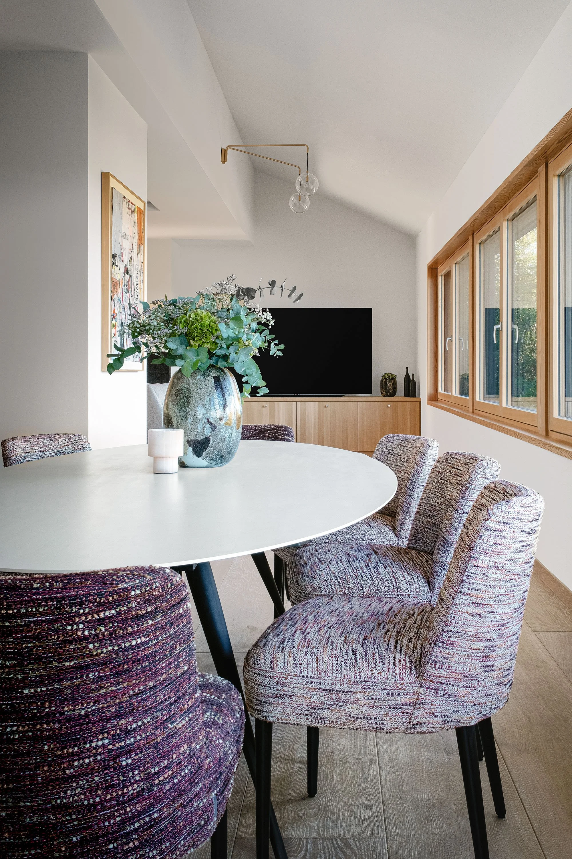 Salle à manger avec table ovale blanche, six chaises recouvertes de tissu multicolore, vase avec fleurs et plantes vertes au centre, grand mur avec fenêtres en bois, téléviseur et décoration minimalist
