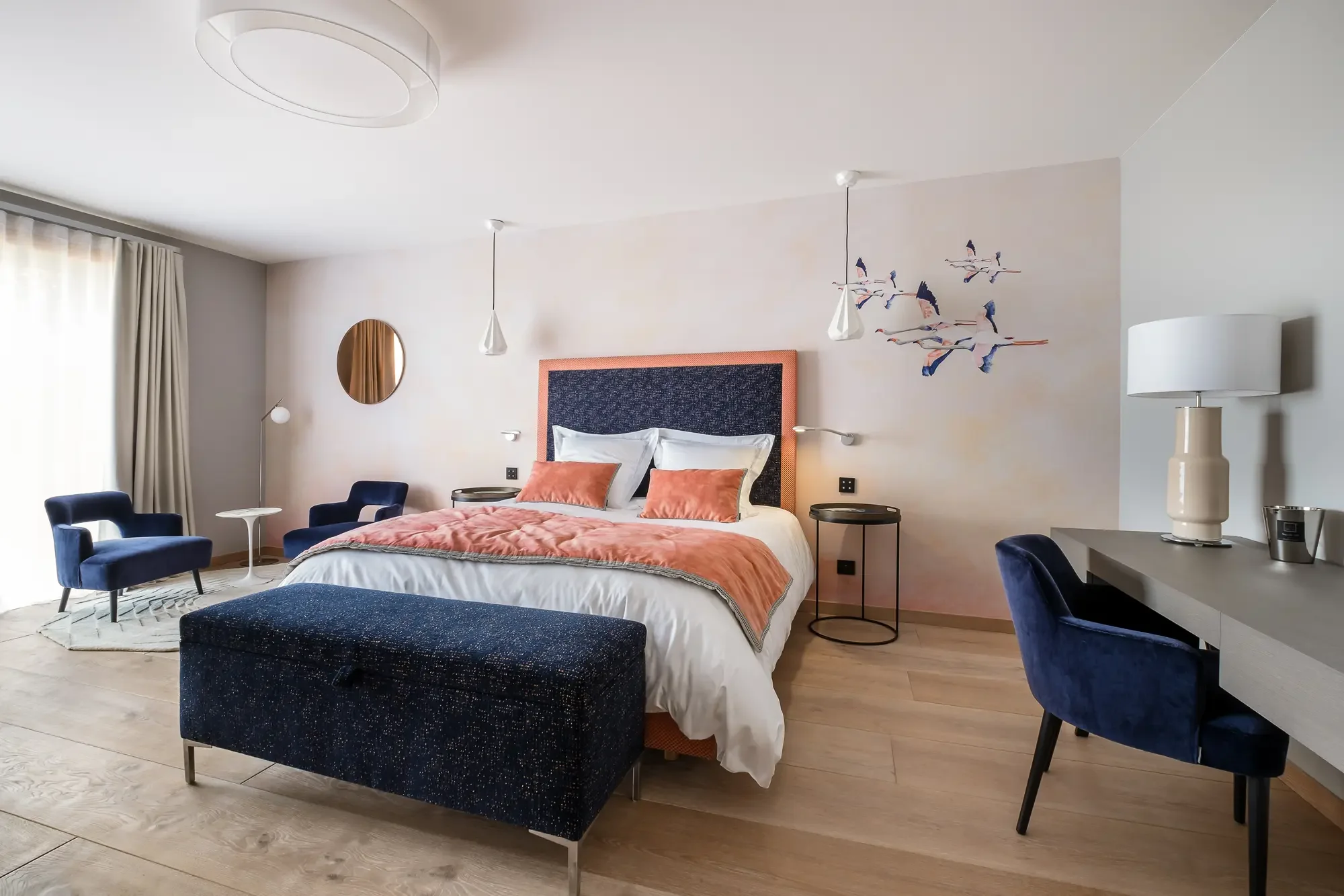 Chambre d'hôtel moderne avec un lit, deux fauteuils bleu foncé, une lampe de chevet, un tapis, un mur décoratif, une grande fenêtre avec rideaux, un bureau avec une lampe et une cafetière.