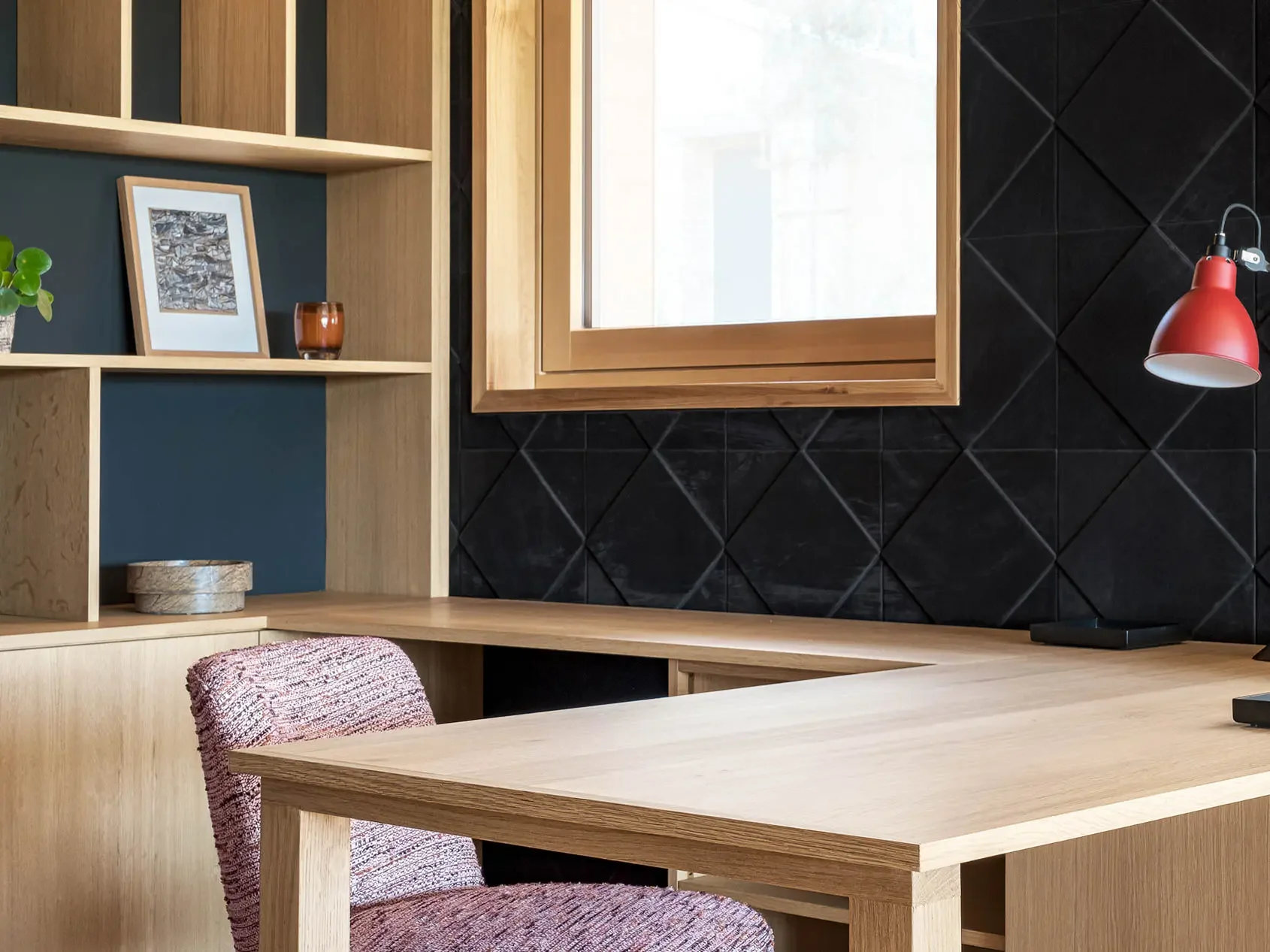 Bureau en bois clair avec une chaise en tissu rose, une étagère murale en bois, une fenêtre avec cadre en bois, un mur noir en diagonale, et une lampe rouge.