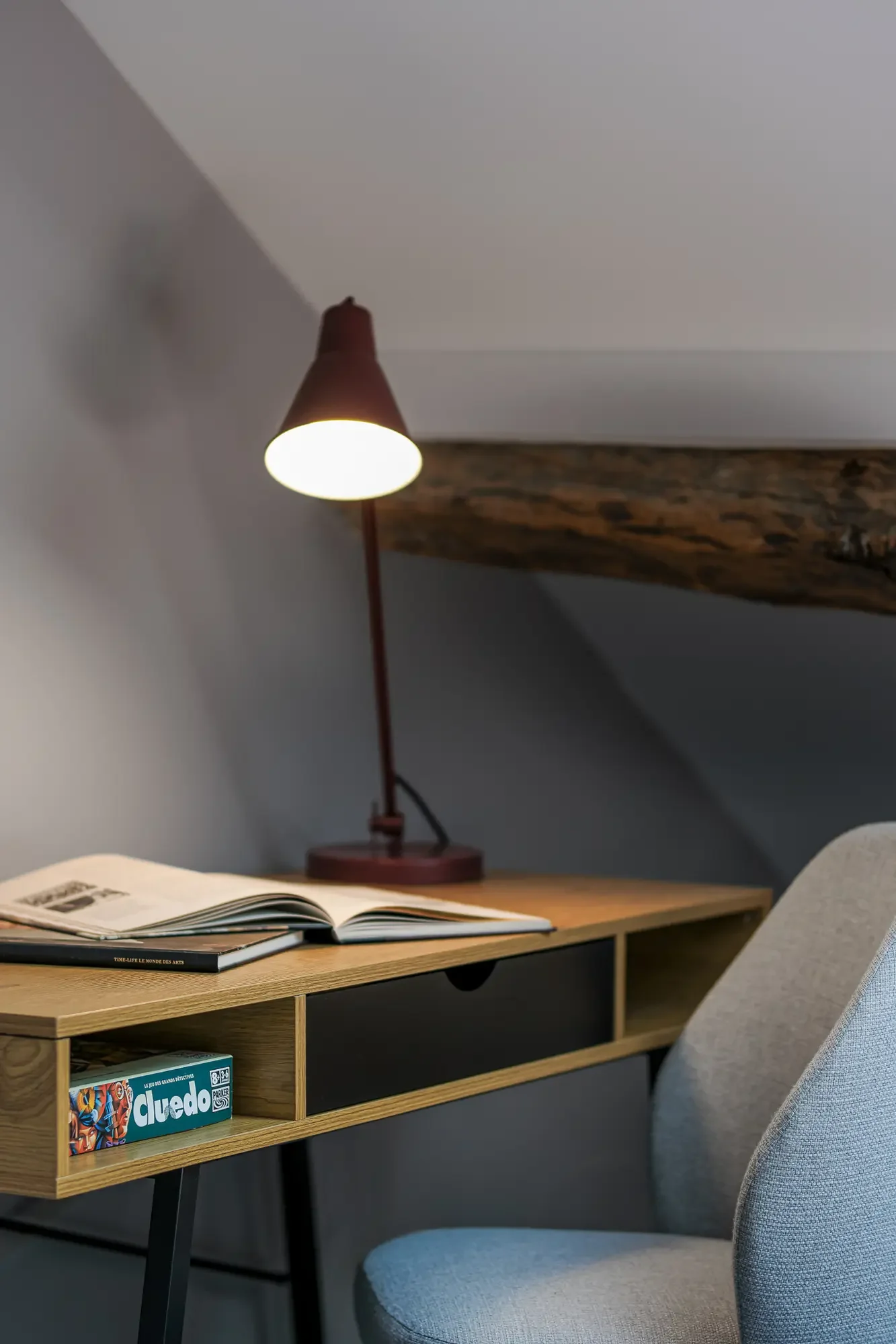 Un bureau avec une lampe de lecture, un livre ouvert, une boîte de jeu de société 'duo', une chaise grise, un mur blanc, et une poutre en bois dans une pièce à sous pente.