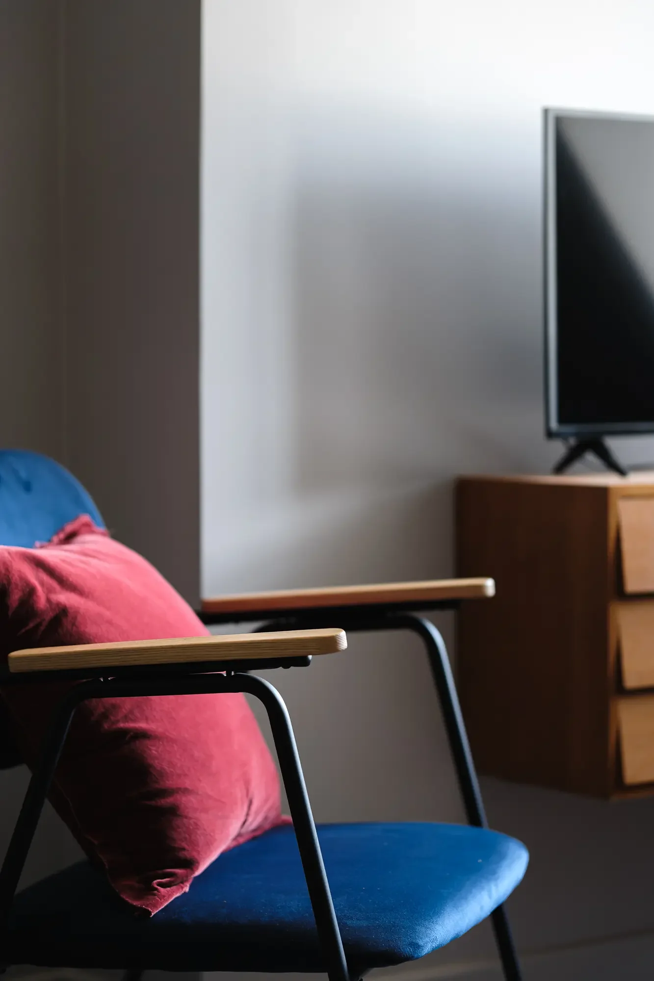 Un fauteuil bleu avec un coussin rouge, dans une pièce lumineuse, avec une télévision à l'arrière et un meuble en bois avec tiroirs.