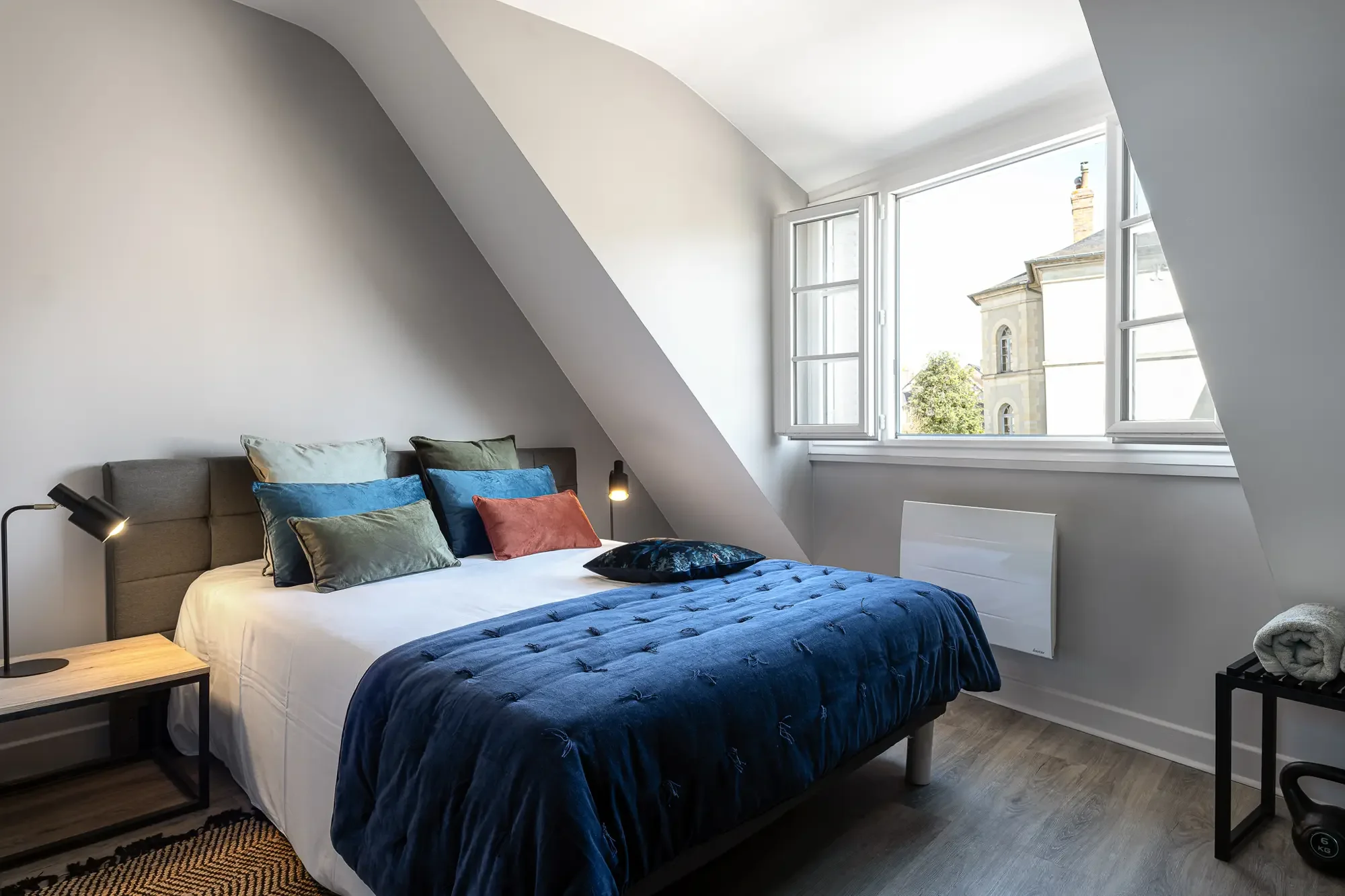 Chambre à coucher moderne avec lit, plusieurs coussins colorés, lampes de chevet, et fenêtre laissant entrer la lumière naturelle.