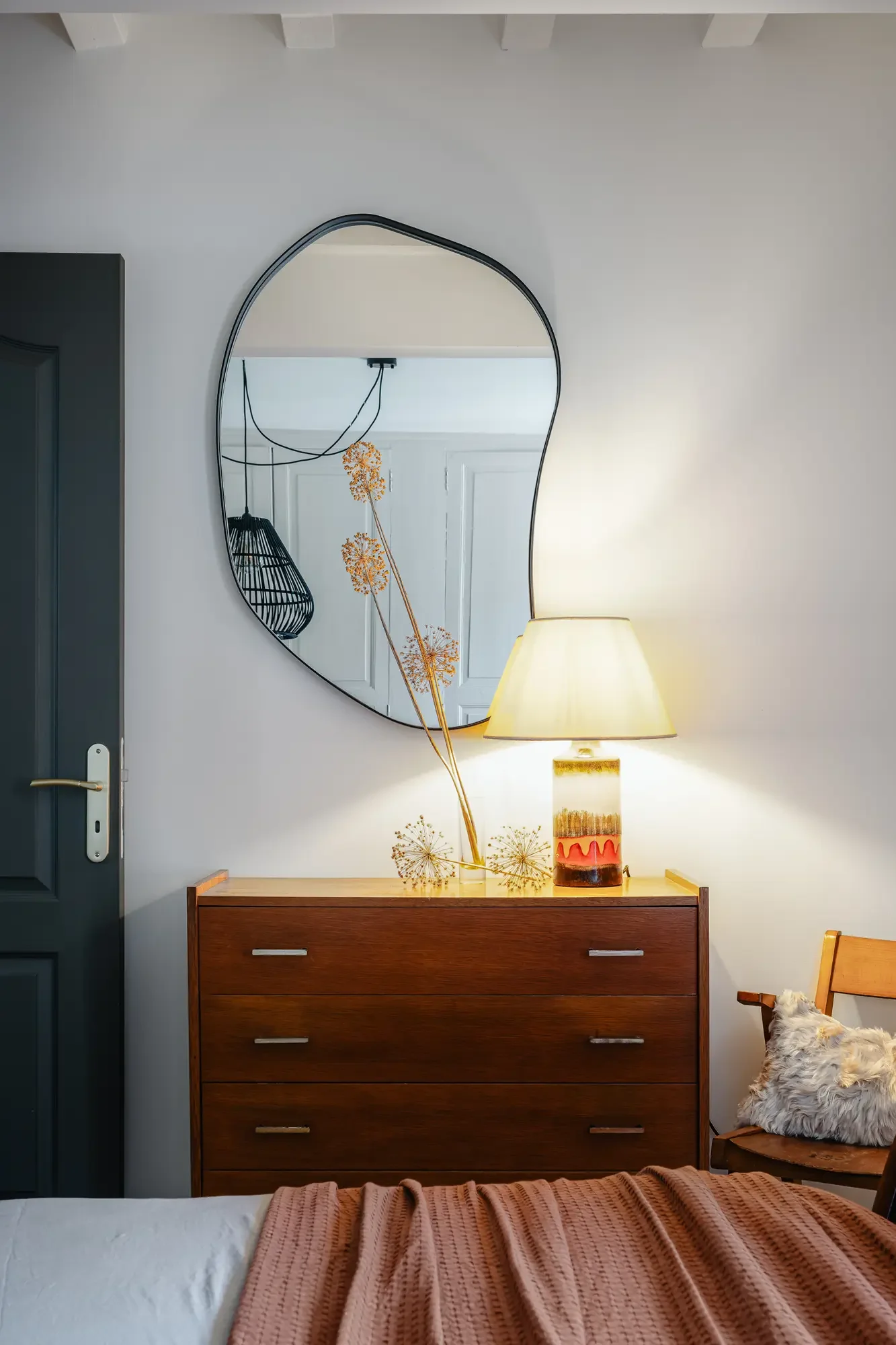 Un miroir design accroché à un mur blanc au-dessus d'une commode en bois, avec une lampe et des décorations florales en dessous.