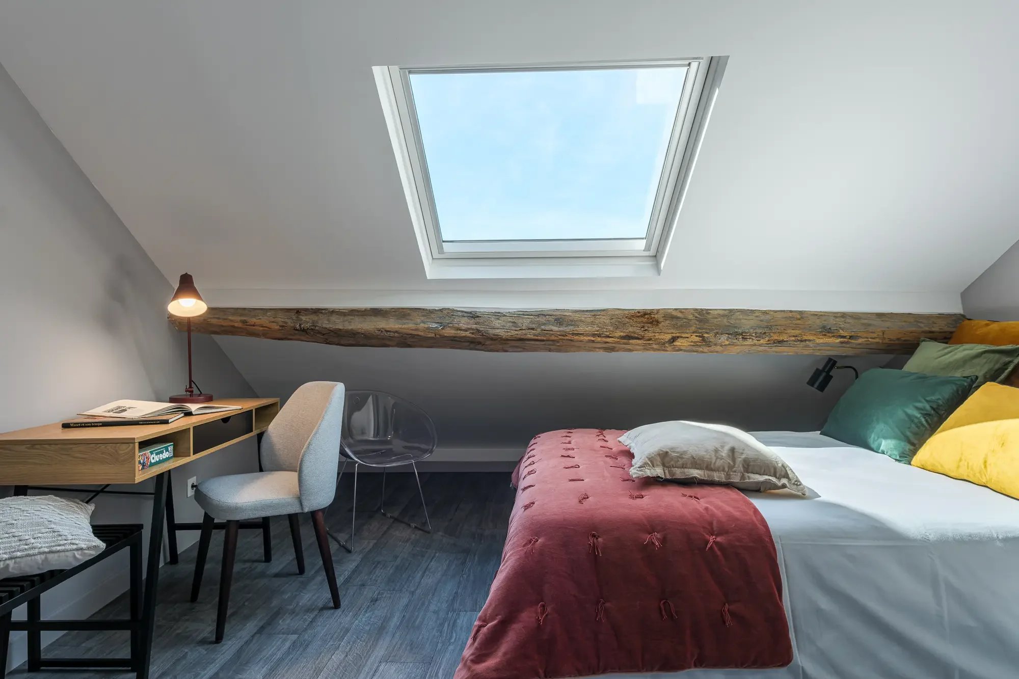 Chambre sous un toit en pente avec un velux au plafond, un lit avec plusieurs coussins, un bureau avec une lampe, une chaise en plastique transparent et une autre en tissu, ainsi qu'une poutre en bois apparente.