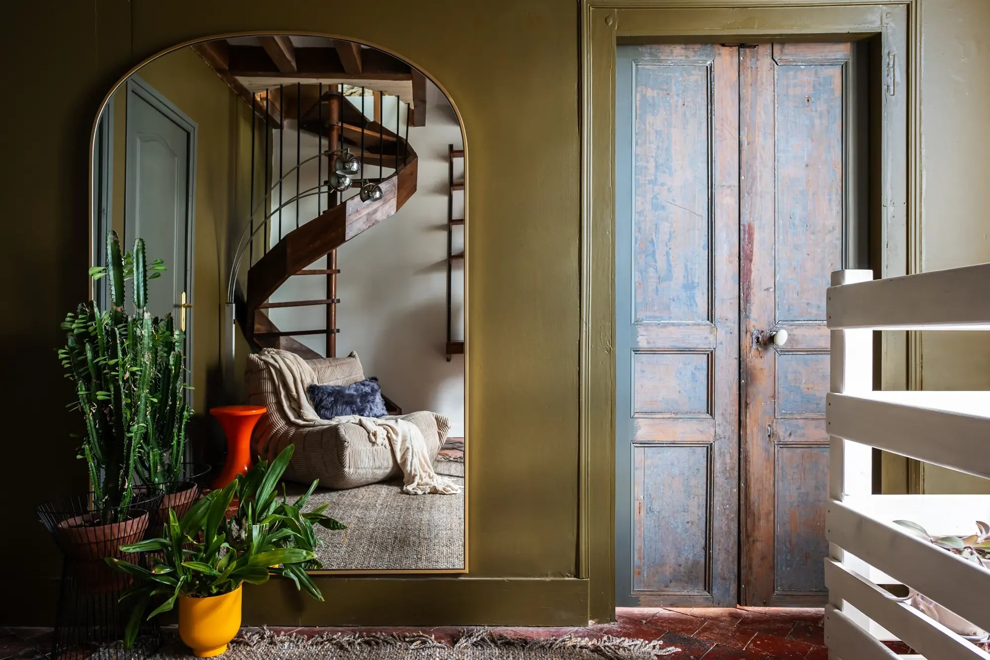 Vue intérieure d'un salon avec un fauteuil, des plantes en pot, un miroir, un escalier en colimaçon en bois et une porte en bois patinée.