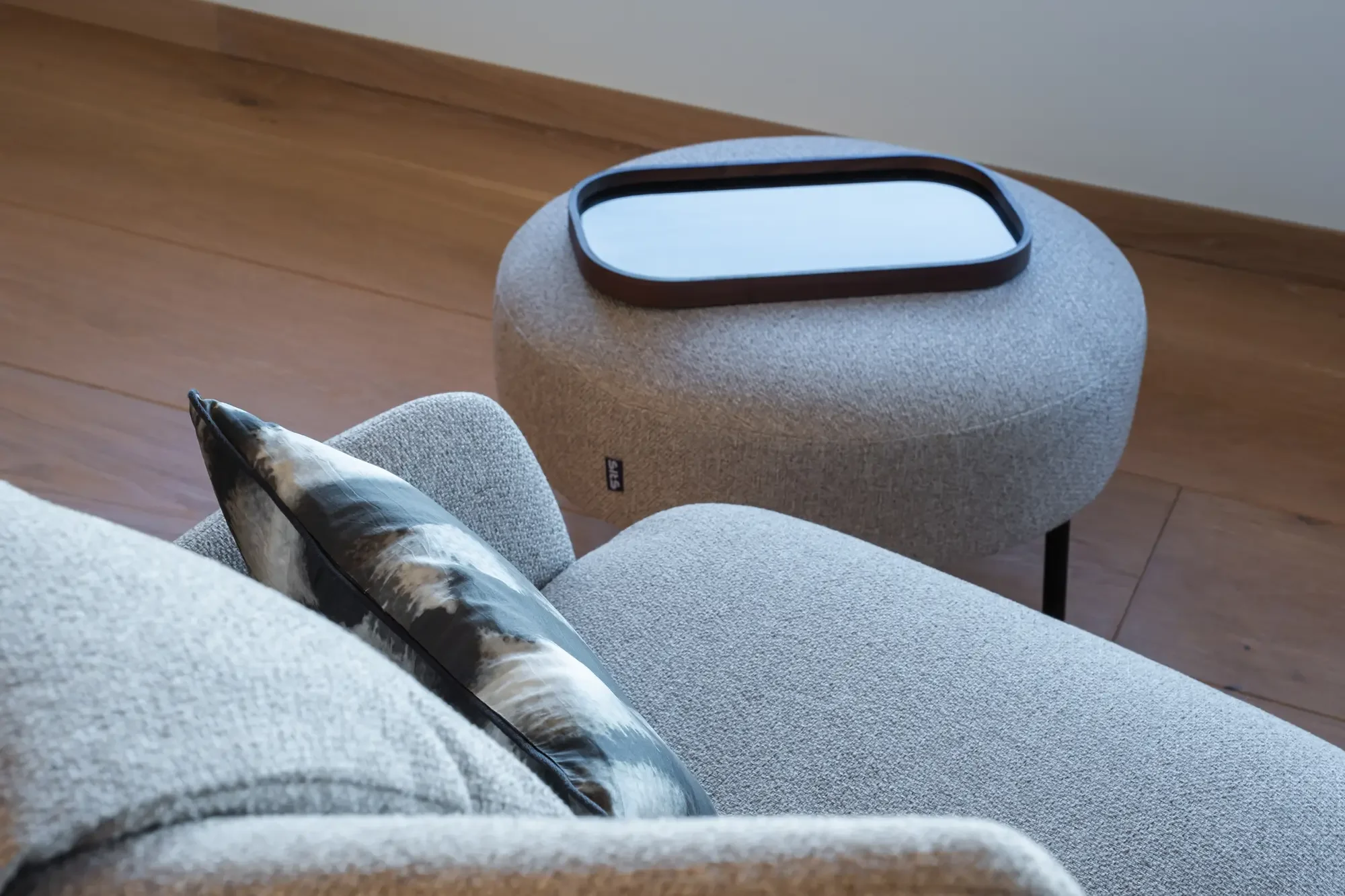 Un fauteuil gris clair avec un coussin décoratif et une table d'appoint ovale en tissu gris, avec un miroir carré posé dessus, dans un intérieur moderne.