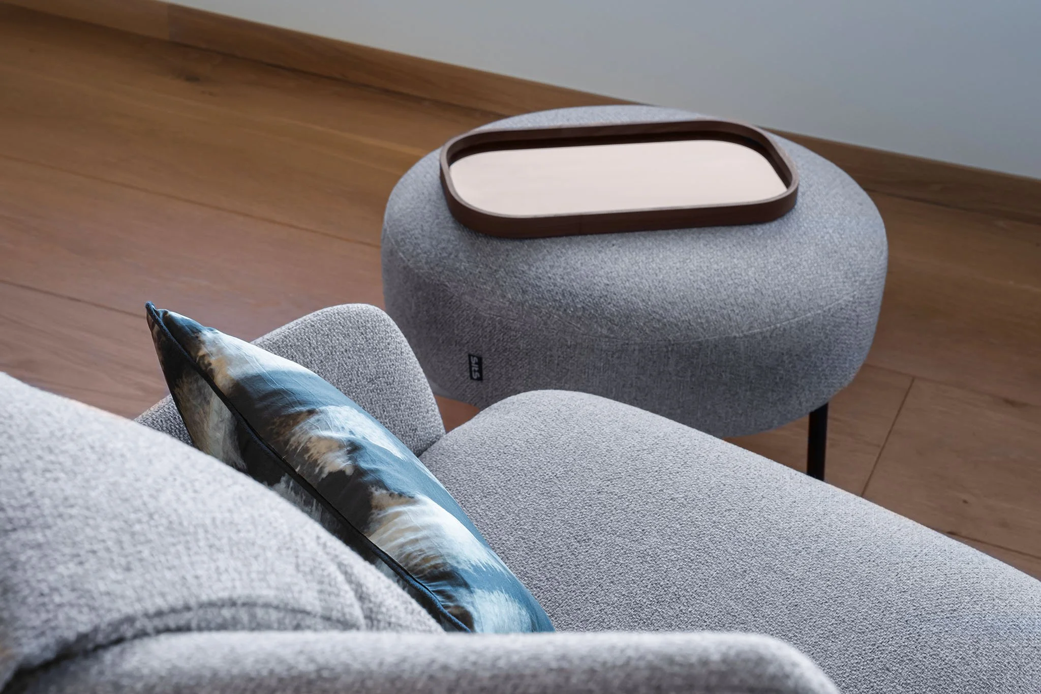 Coin d'un salon moderne avec un canapé gris, un coussin avec un motif de plage ou de mer, une table basse ovale en tissu gris foncé, et un sol en bois clair.