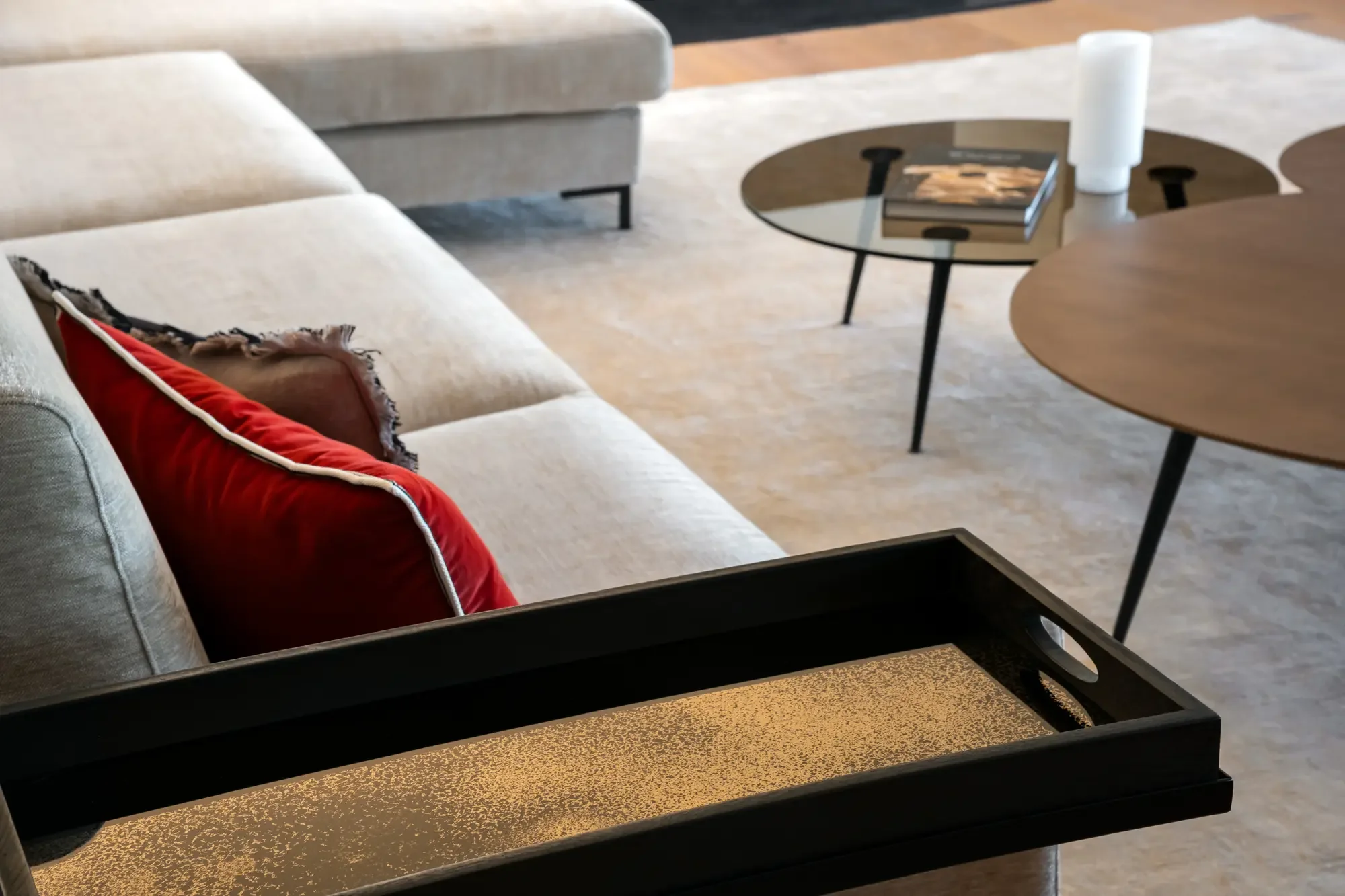 Salon avec canapé beige, coussins rouges et marron, tables basses en bois et verre, tapis beige, et un plateau noir avec un fond doré.