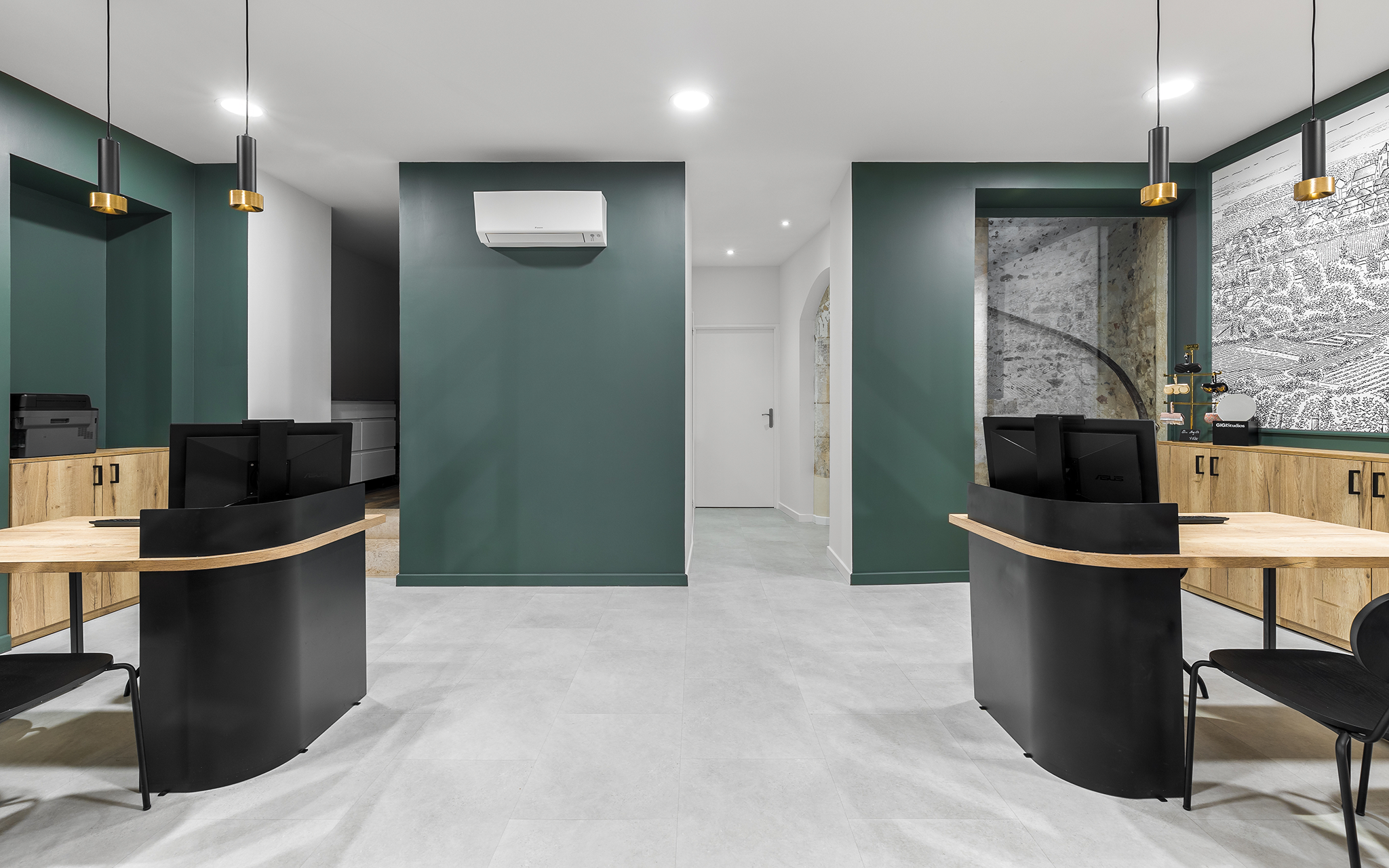 Bureau moderne avec deux postes de travail, murs verts, meubles en bois, décoration élégante, éclairage suspendu, encadrements en béton apparent, espace organisé, environnement professionnel.