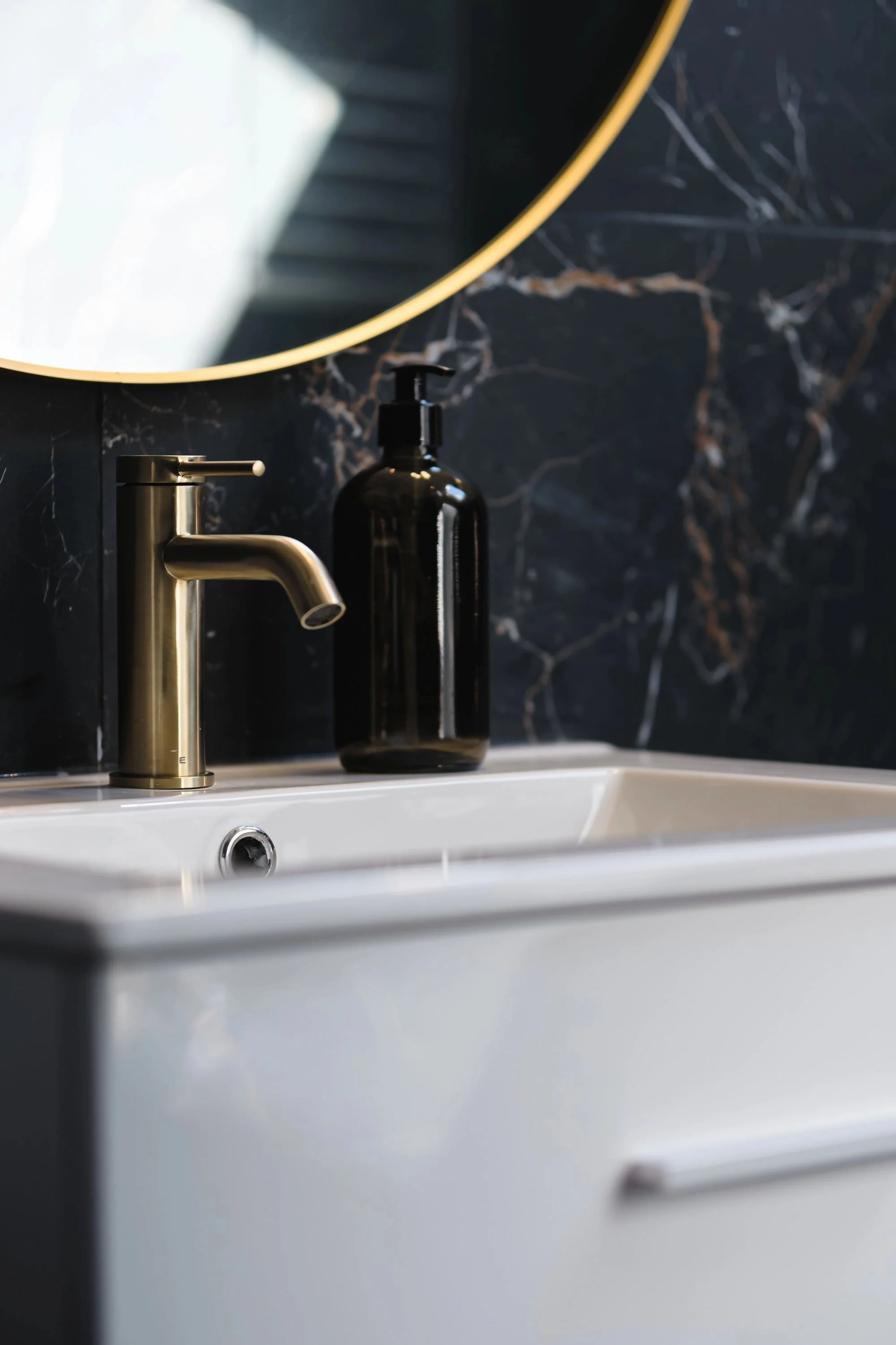Lavabo blanc avec robinet en métal argenté, bouteille de savon noire sur un comptoir noir marbré, miroir rond avec bord doré au-dessus.