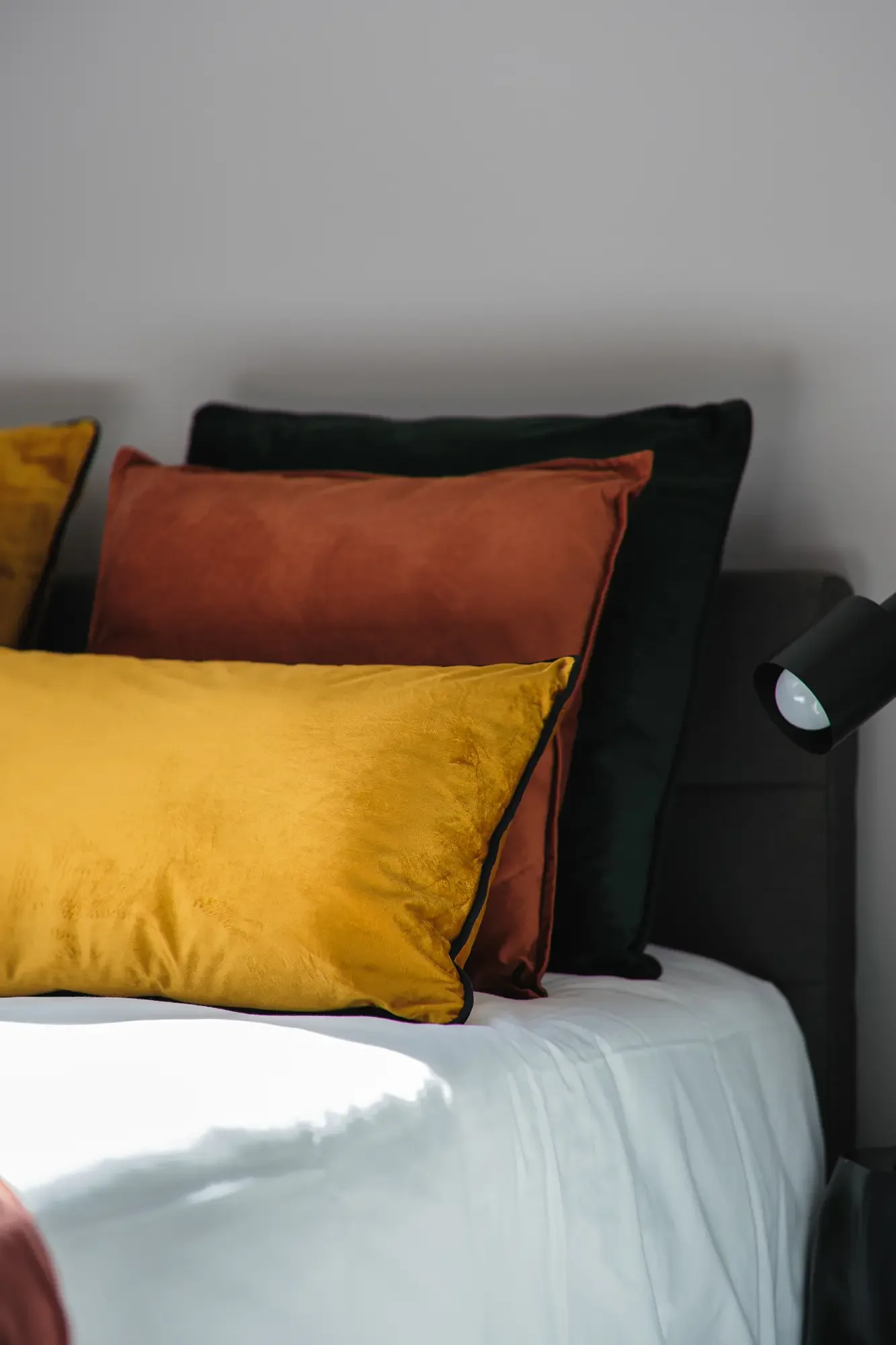 Coussins de couleur jaune, rouille et noir sur un lit avec une tête de lit en tissu noir, et une lampe de chevet noire à côté.