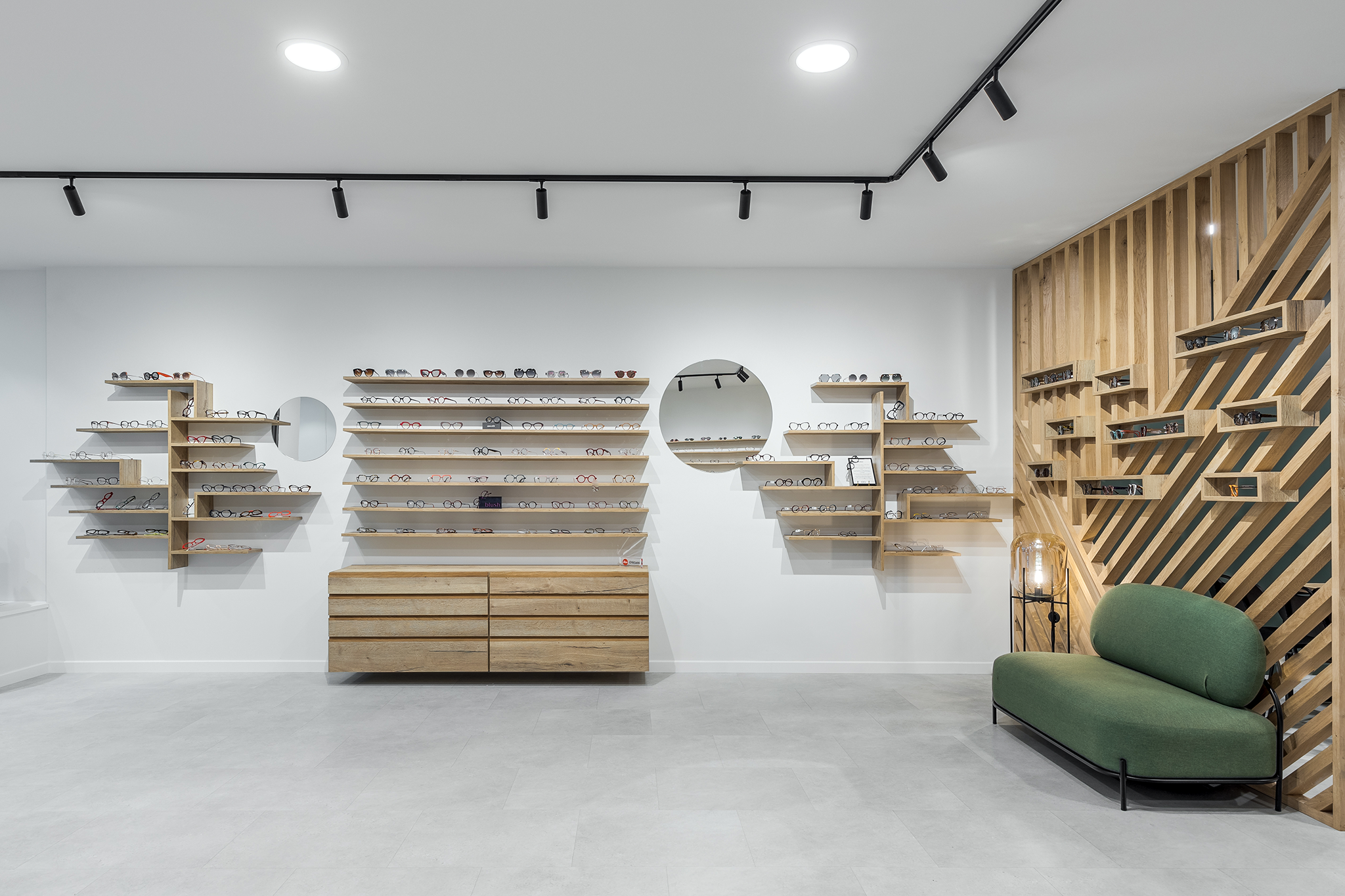 Vitrine d'une boutique de lunettes avec plusieurs étagères en bois affichant des montures de différentes formes et couleurs, miroir rond sur le mur, et coin salon avec canapé vert et lampe de sol.