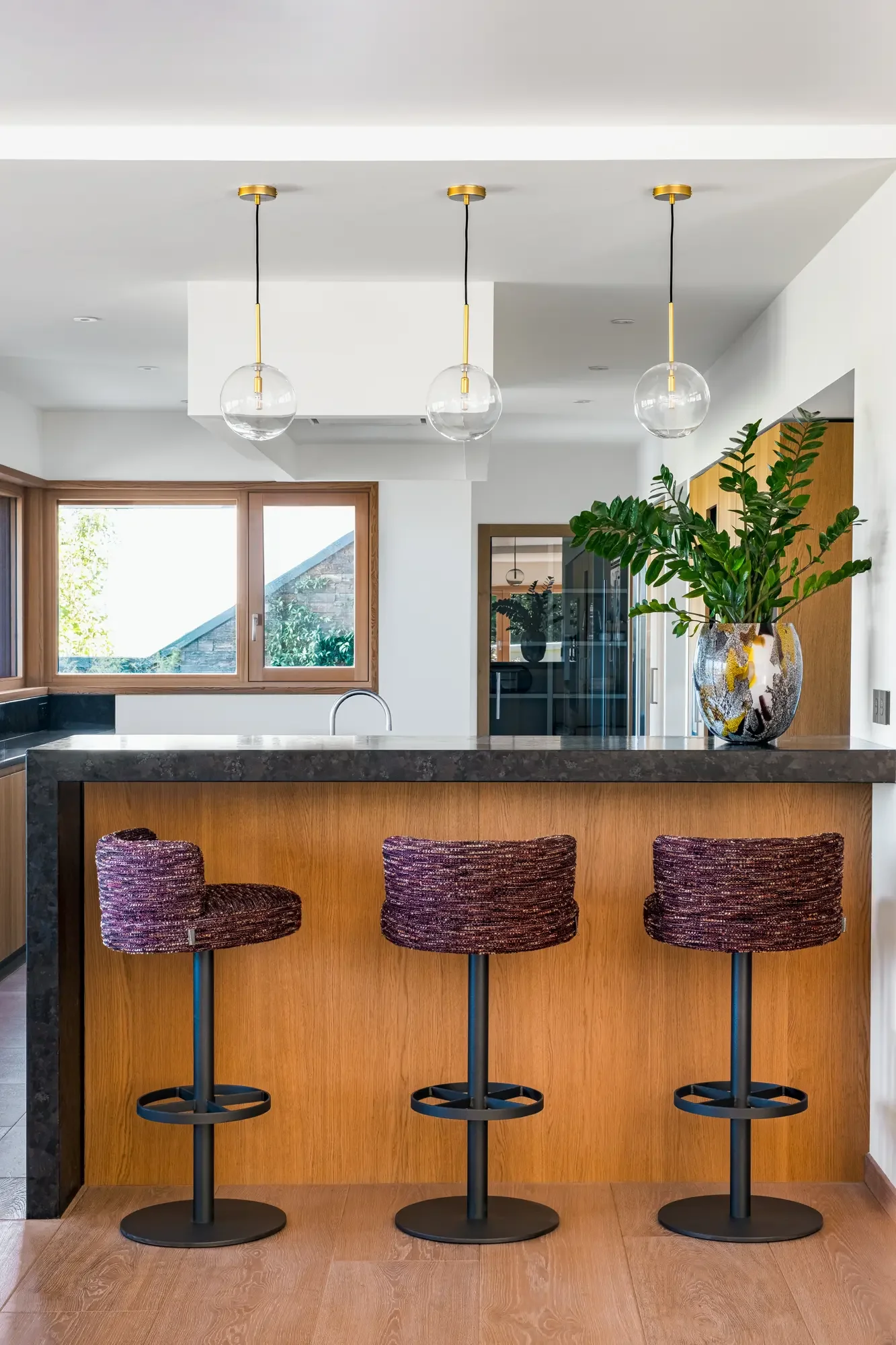 Une cuisine moderne avec un comptoir en pierre, trois chaises de bar rembourrées, un grand vase avec une plante verte, et trois lampes suspendues à abat-jour en verre.