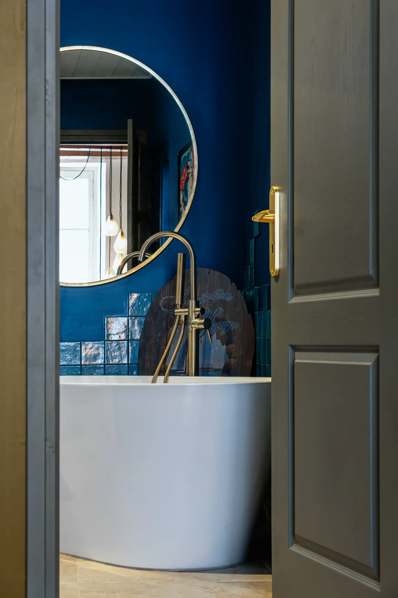 Vue partielle d'une salle de bain avec baignoire autoportante, miroir rond, murs peints en bleu, et fenêtres laissant entrer la lumière naturelle.